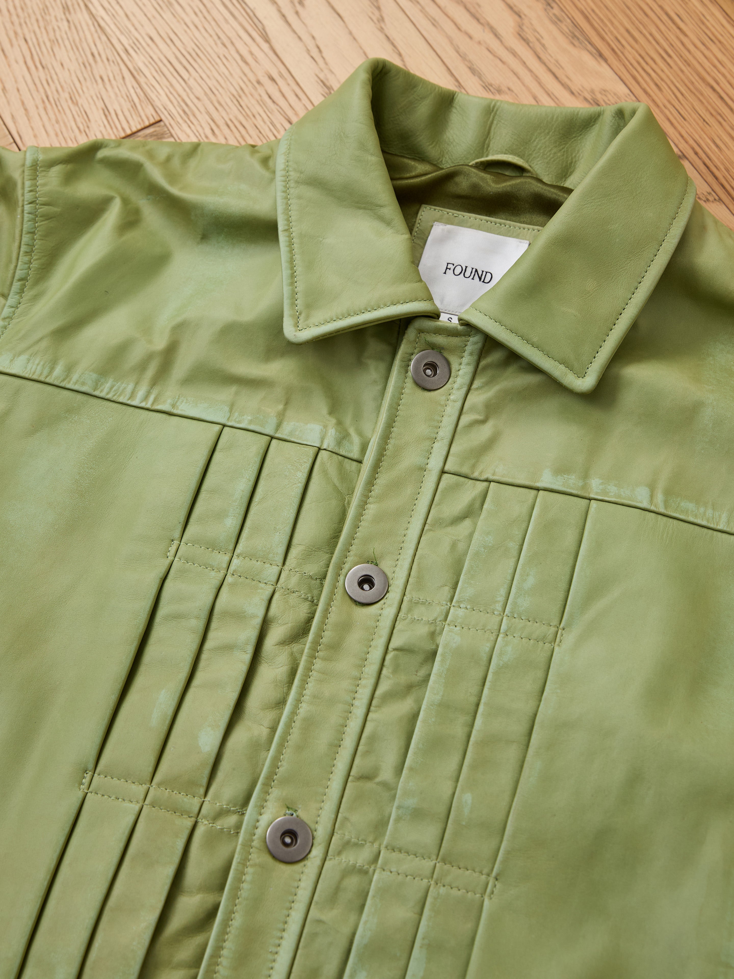 The FOUND Trucker Leather Jacket features washed green leather, vertical stitched details, dark buttons, and a relaxed unisex fit, finished with a signature white FOUND label inside. Displayed on a wooden floor.