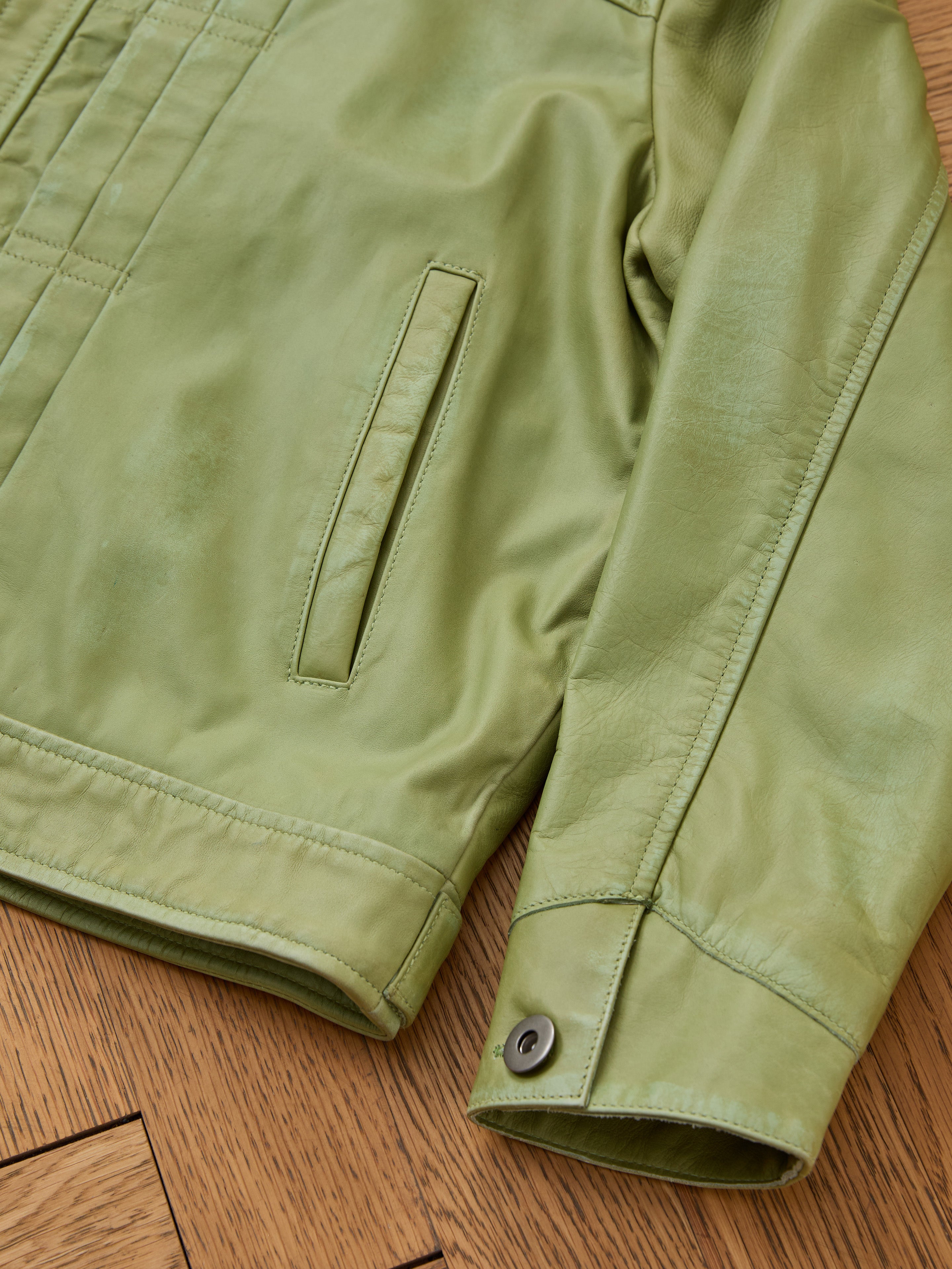 FOUND Trucker Leather Jacket in washed green, unisex style featuring a side pocket and buttoned cuffs, displayed flat on a wooden floor.