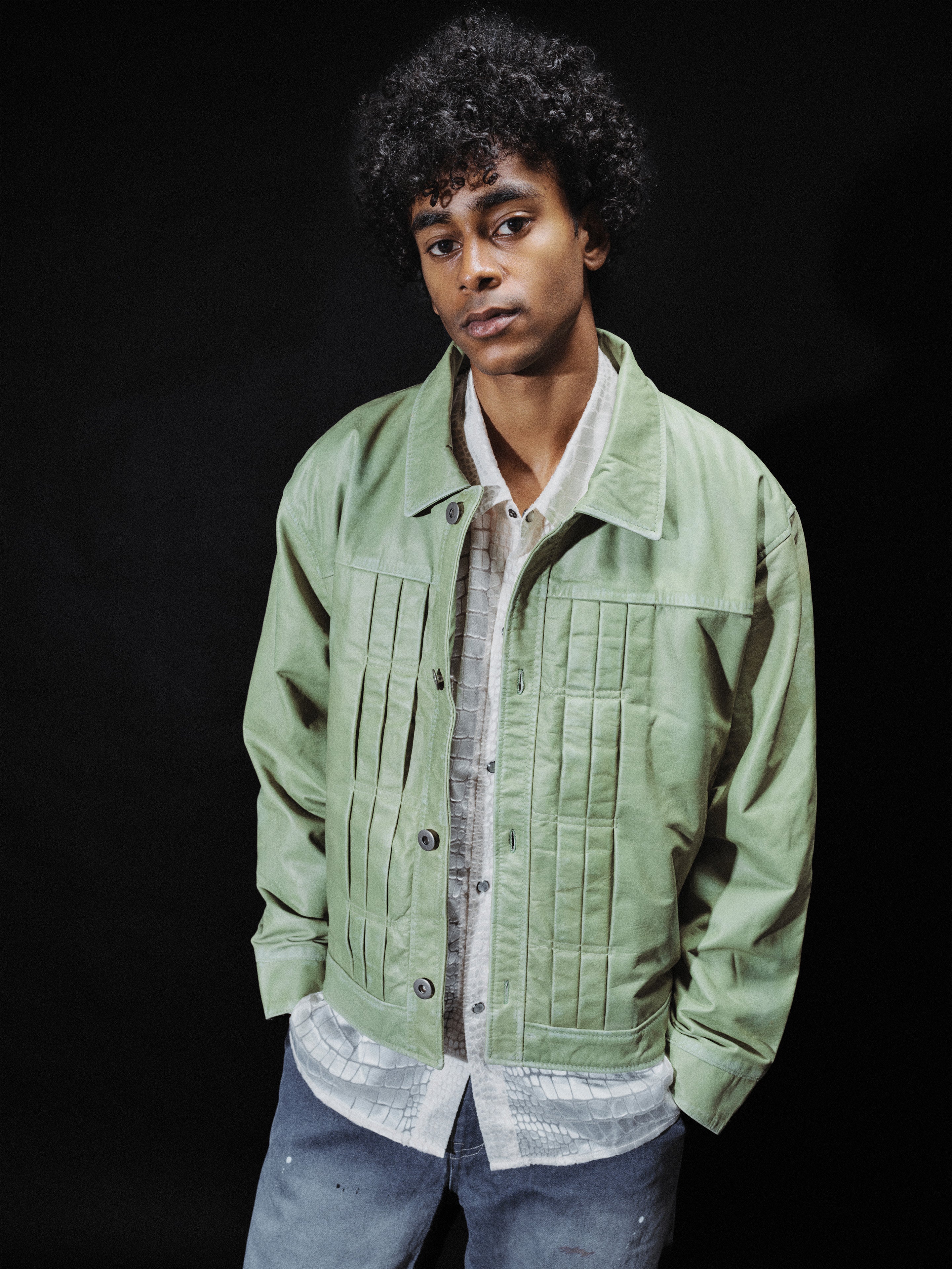 A person with curly hair wears the FOUND Trucker Leather Jacket in washed green, featuring a relaxed fit, layered over a patterned shirt and blue jeans, standing against a plain dark background.