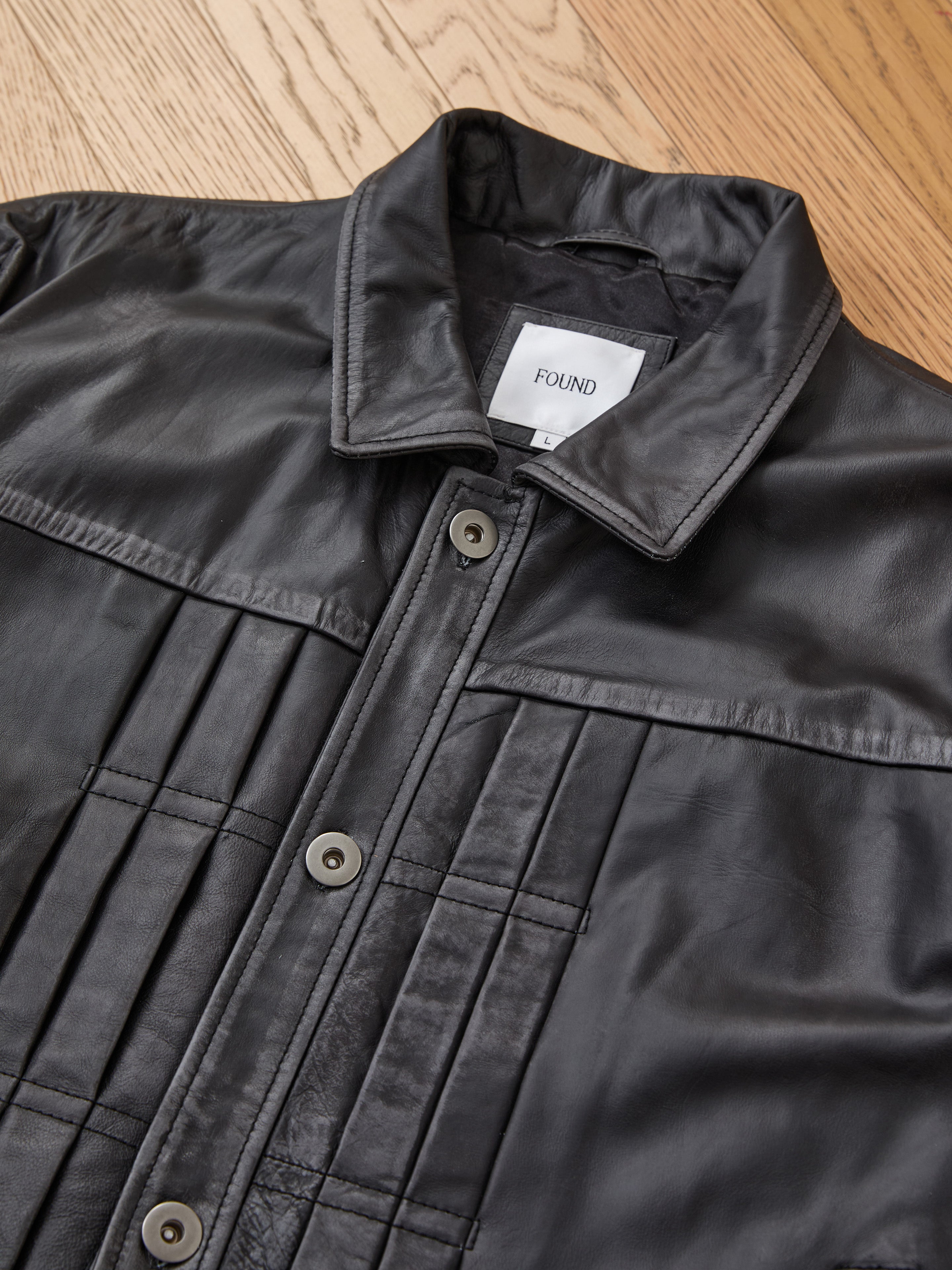 FOUND Trucker Leather Jacket in black leather with vertical stitching and button closure, featuring a visible FOUND label, displayed on a wooden floor.