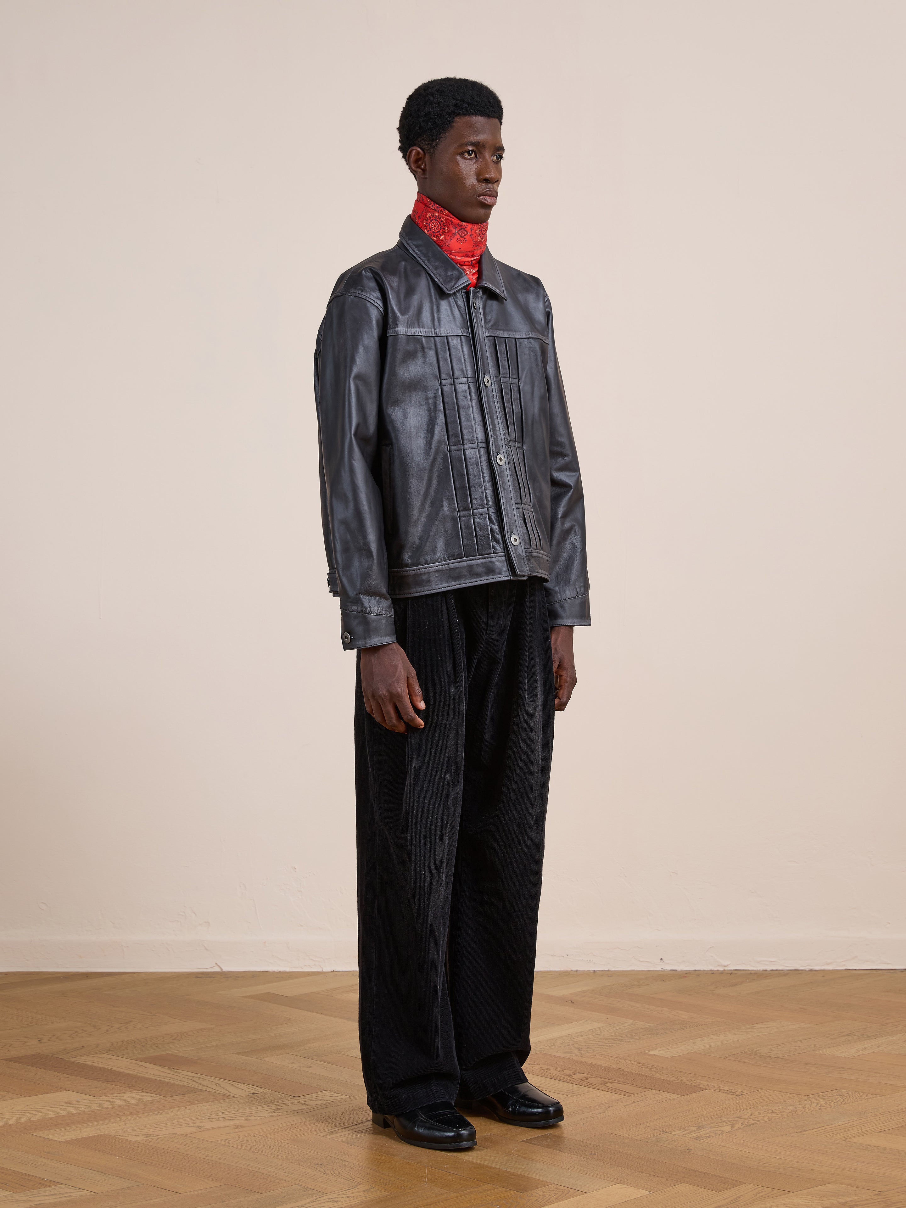 Indoors, a person models the FOUND Trucker Leather Jacket with black pants, black shoes, and a red bandana around their neck, standing against a plain light wall and wooden floor.