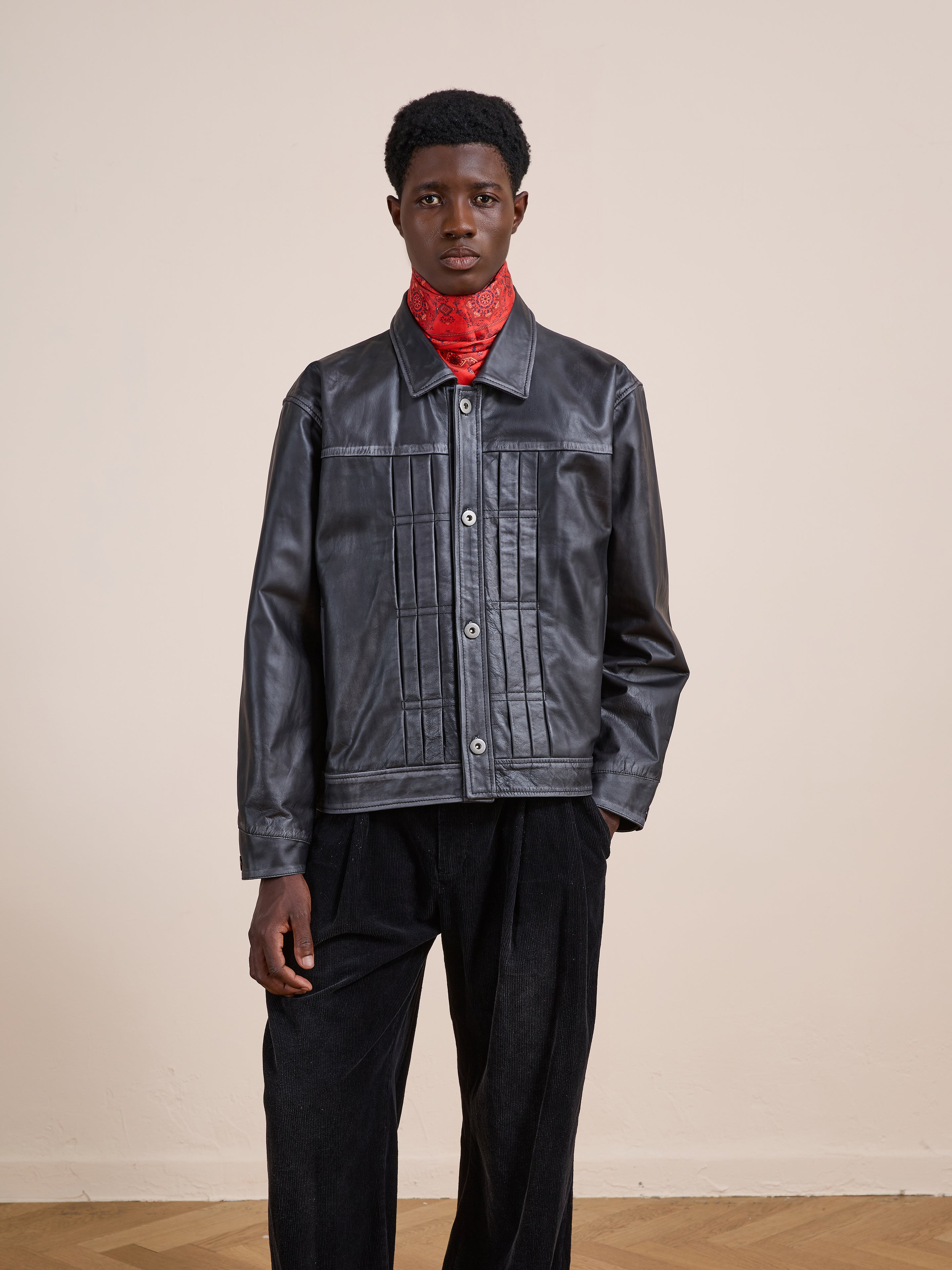 A person stands against a plain background wearing the FOUND Trucker Leather Jacket, paired with a red patterned scarf and black pants.