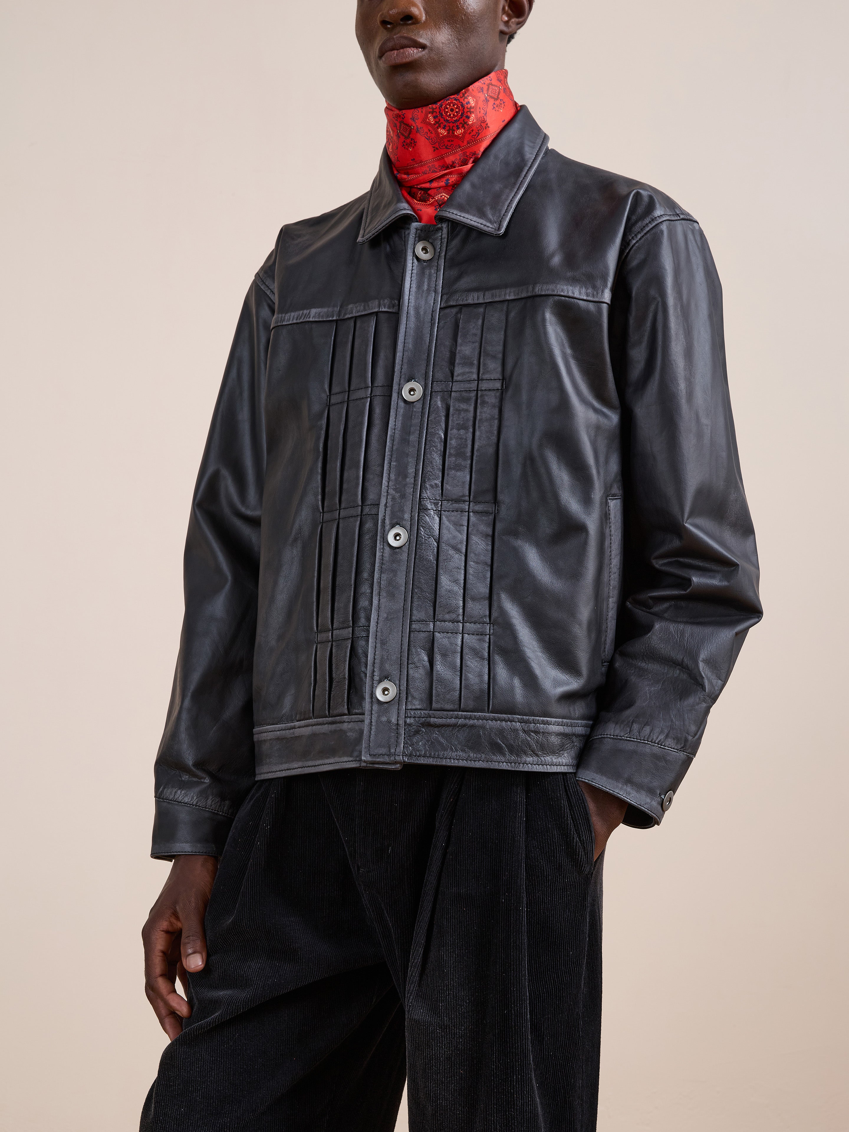 A person wears the FOUND Trucker Leather Jacket in black with vertical stitching, styled with black pants and a red patterned neck scarf.