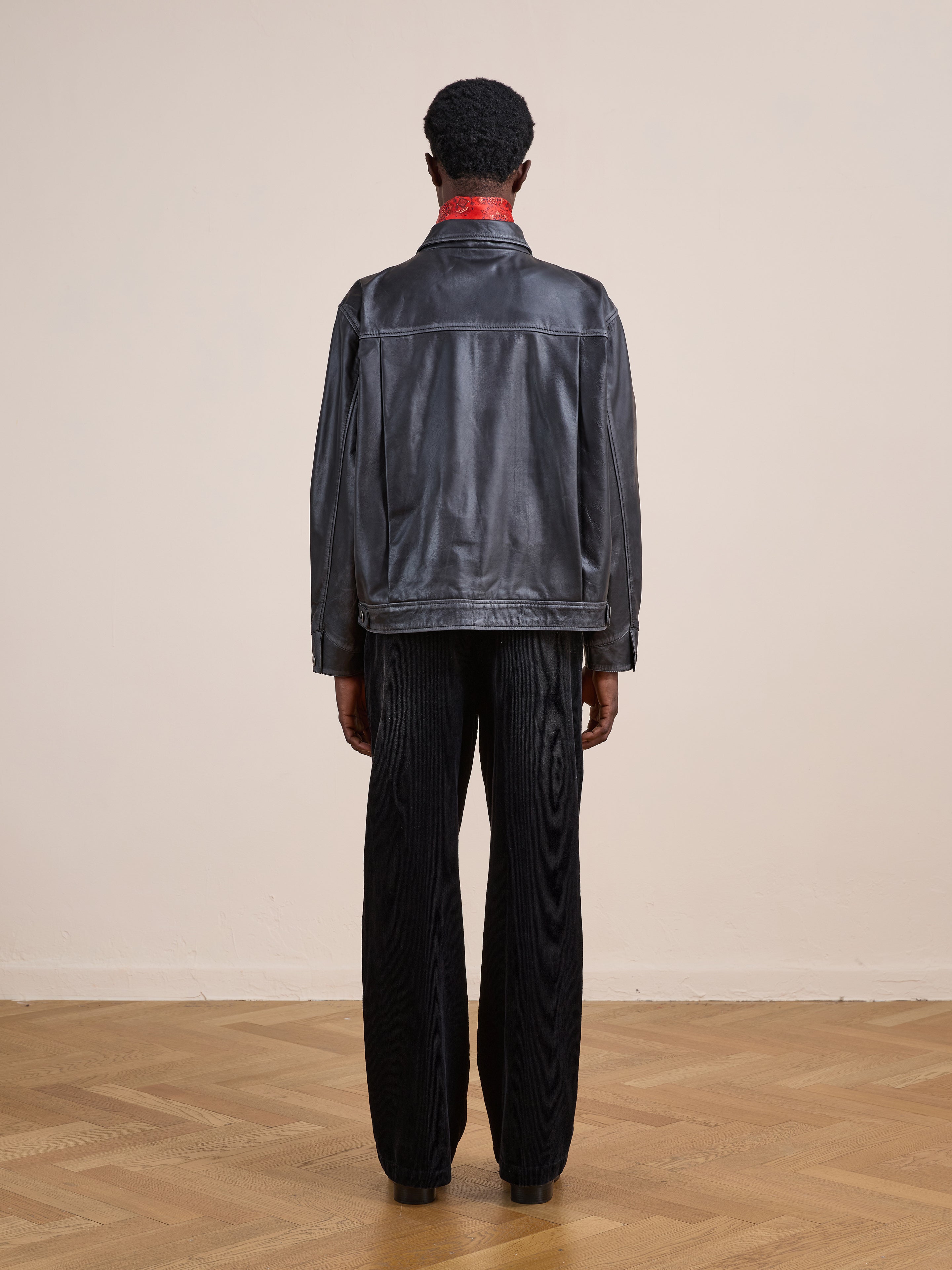 A person stands indoors facing away, wearing the FOUND Trucker Leather Jacket, black pants, and black shoes on a wooden floor against a plain beige wall.