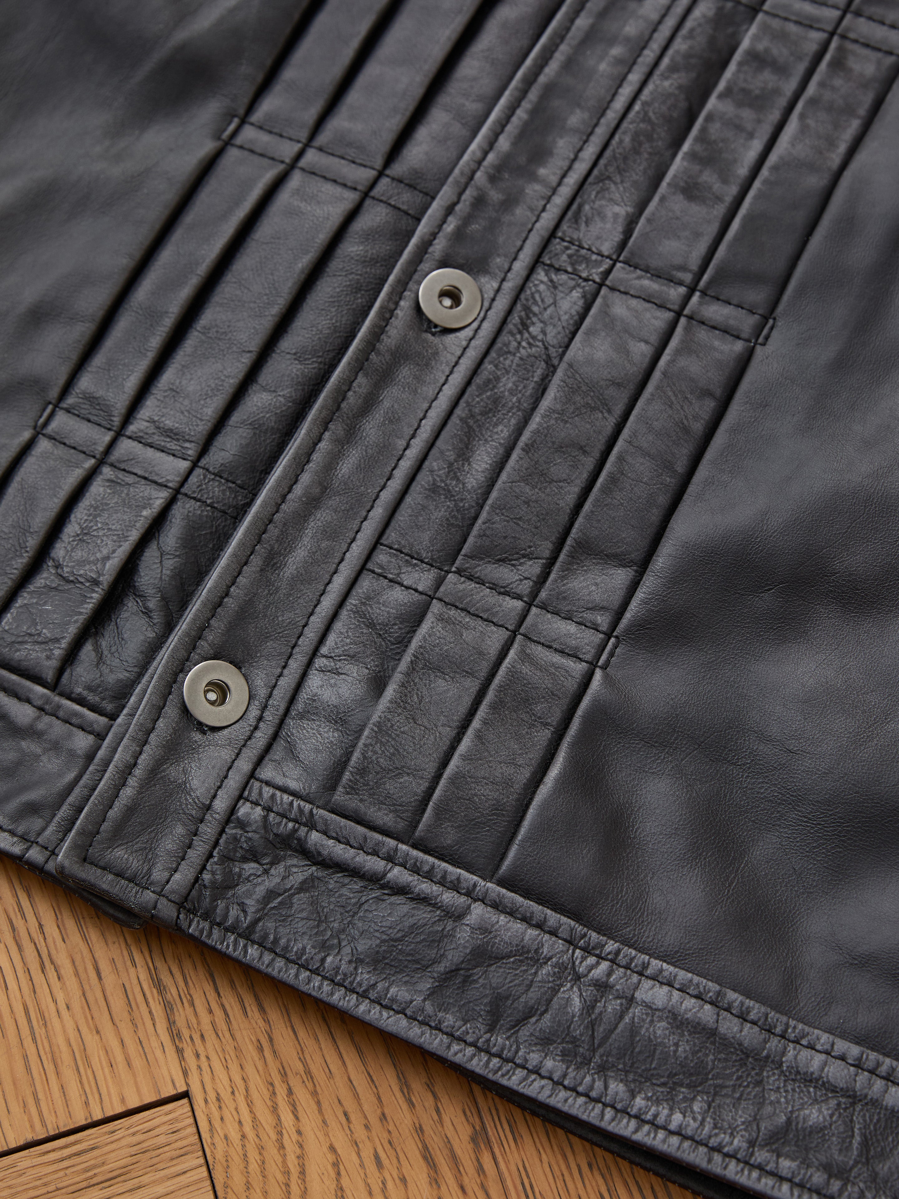 Close-up of the FOUND Trucker Leather Jacket, showcasing its vintage black leather, visible stitching, and metal snap buttons, laid flat on a wooden surface.