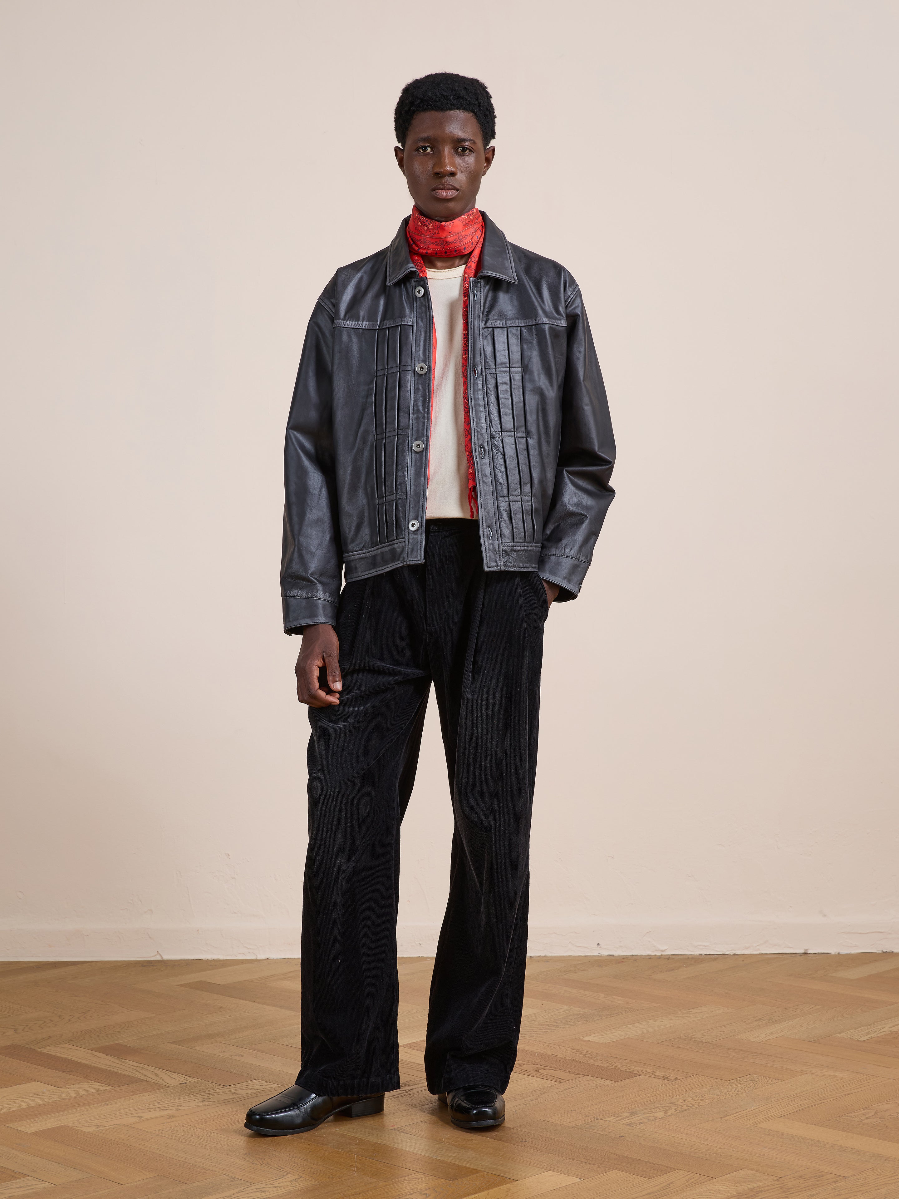 A person stands indoors against a light wall and wooden floor, wearing the FOUND Trucker Leather Jacket, black pants, black shoes, a white shirt, and a red scarf.