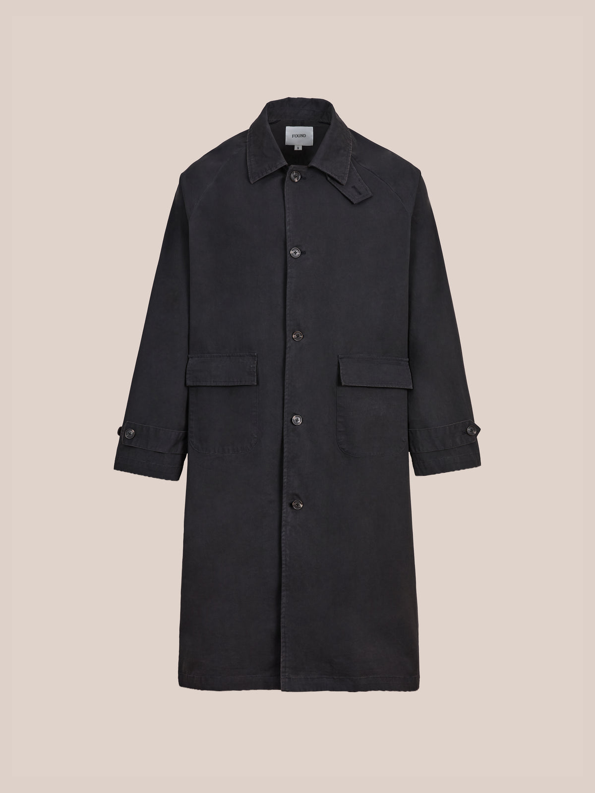The FOUND Twill Work Trench Coat is a long black cotton twill trench featuring a collar, front pockets, and detailed cuffs, shown against a plain beige background. With its relaxed fit, this versatile piece offers timeless style for any occasion.