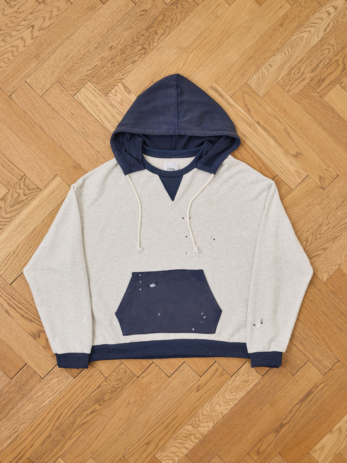 The FOUND Two-Tone Painter Hoodie, featuring a relaxed fit with a navy hood, pocket, and trim accented by subtle paint splatters, is laid flat on a wood herringbone floor.