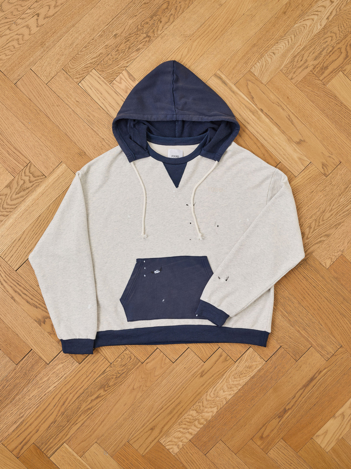 FOUND’s Two-Tone Painter Hoodie, featuring a gray body with navy blue hood, cuffs, and front pocket and subtle vintage-style distressing, is displayed flat on a light wood herringbone floor.