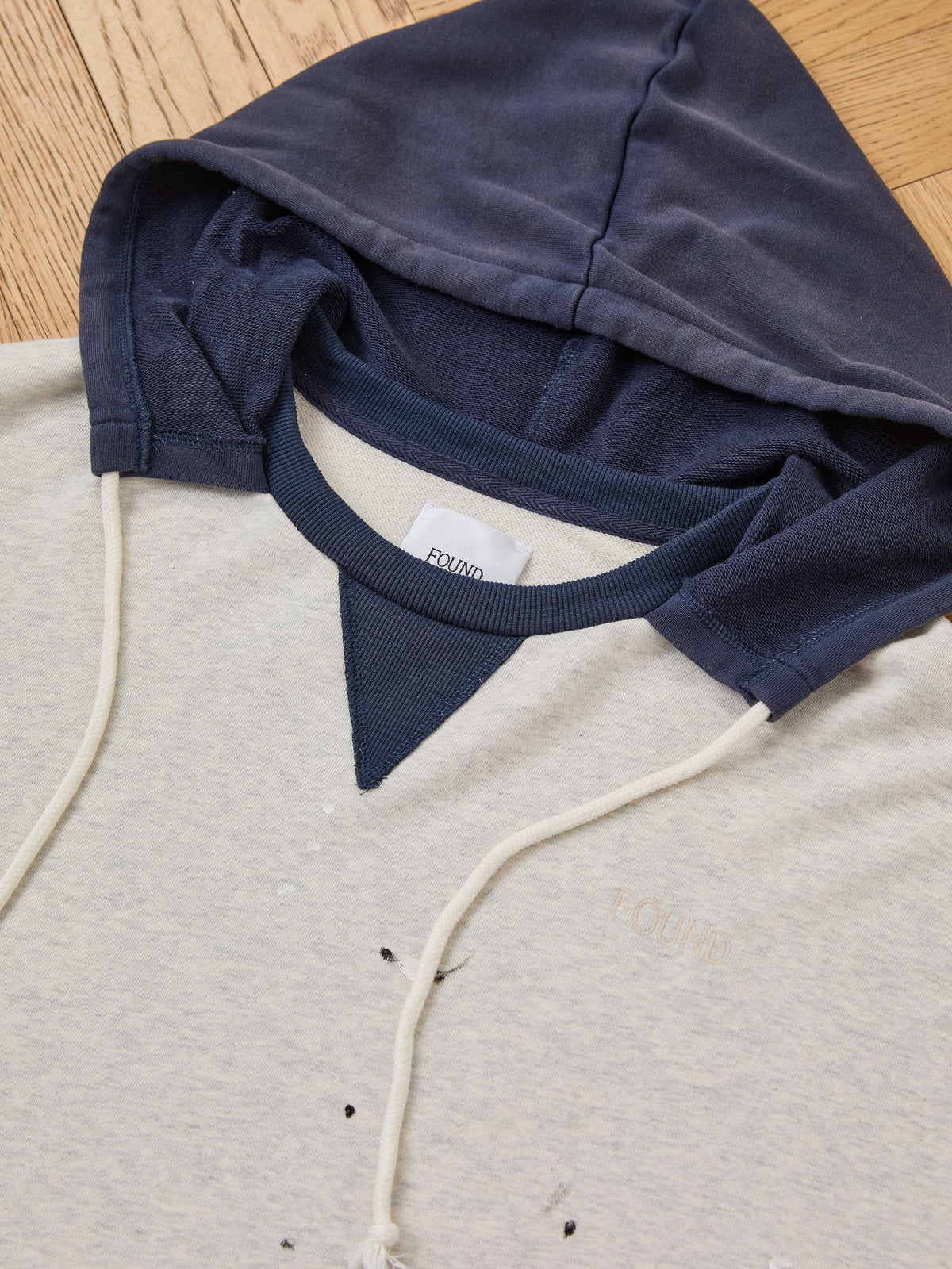 The FOUND Two-Tone Painter Hoodie, featuring a light gray body with a navy blue hood and drawstrings, is displayed on a wooden surface. This relaxed fit hoodie has a small white tag and minor visible stains on the front.
