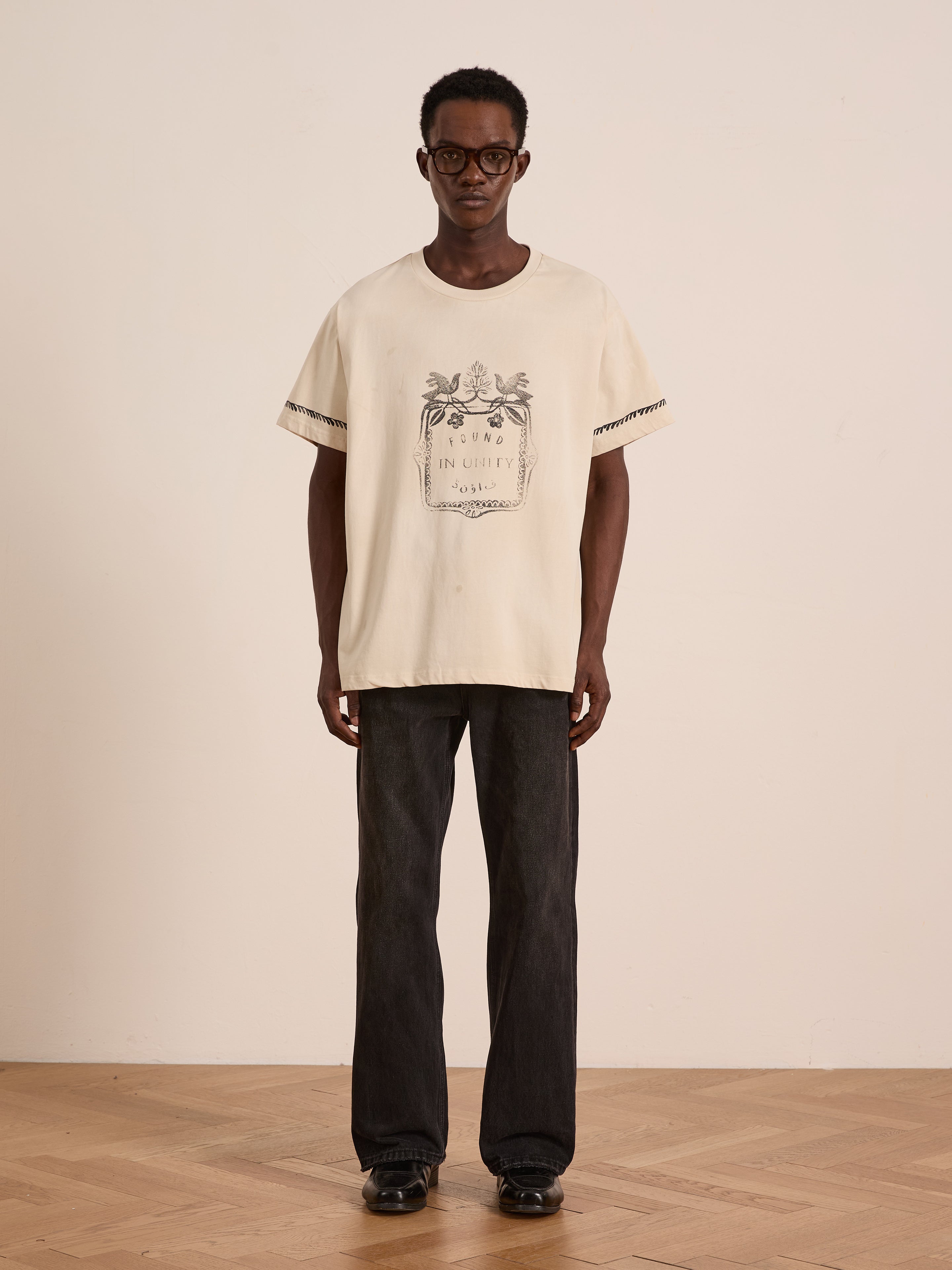 A person stands indoors against a light-colored wall wearing FOUND's Unity Crest Tee, a cream unisex graphic shirt in mid-weight cotton, styled with black glasses, dark straight-leg jeans, and black shoes.
