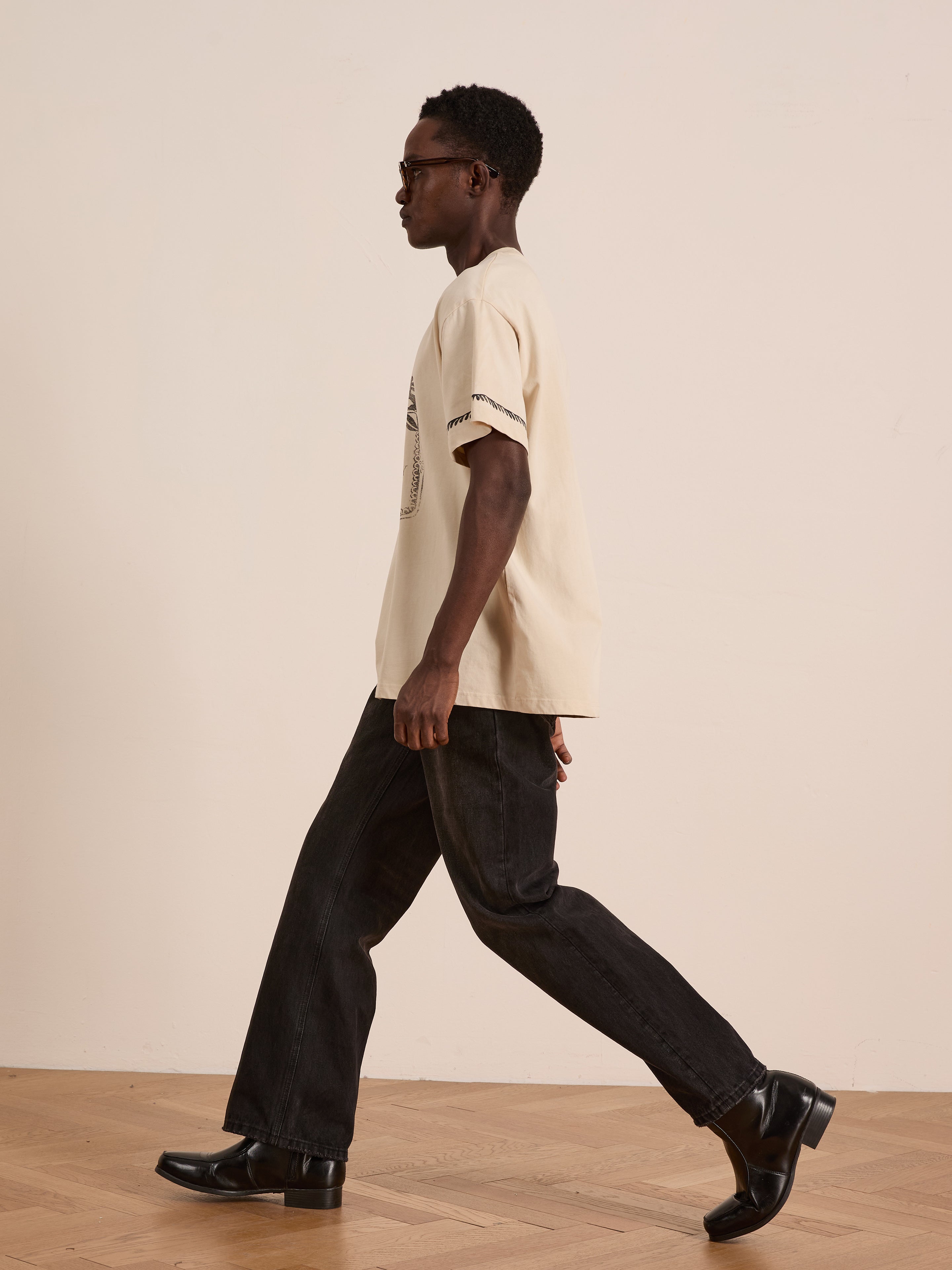 A person wearing the FOUND Unity Crest Tee—a beige, unisex graphic tee made from mid-weight cotton—paired with black jeans and shoes, walks indoors on a wooden floor against a plain light-colored wall.
