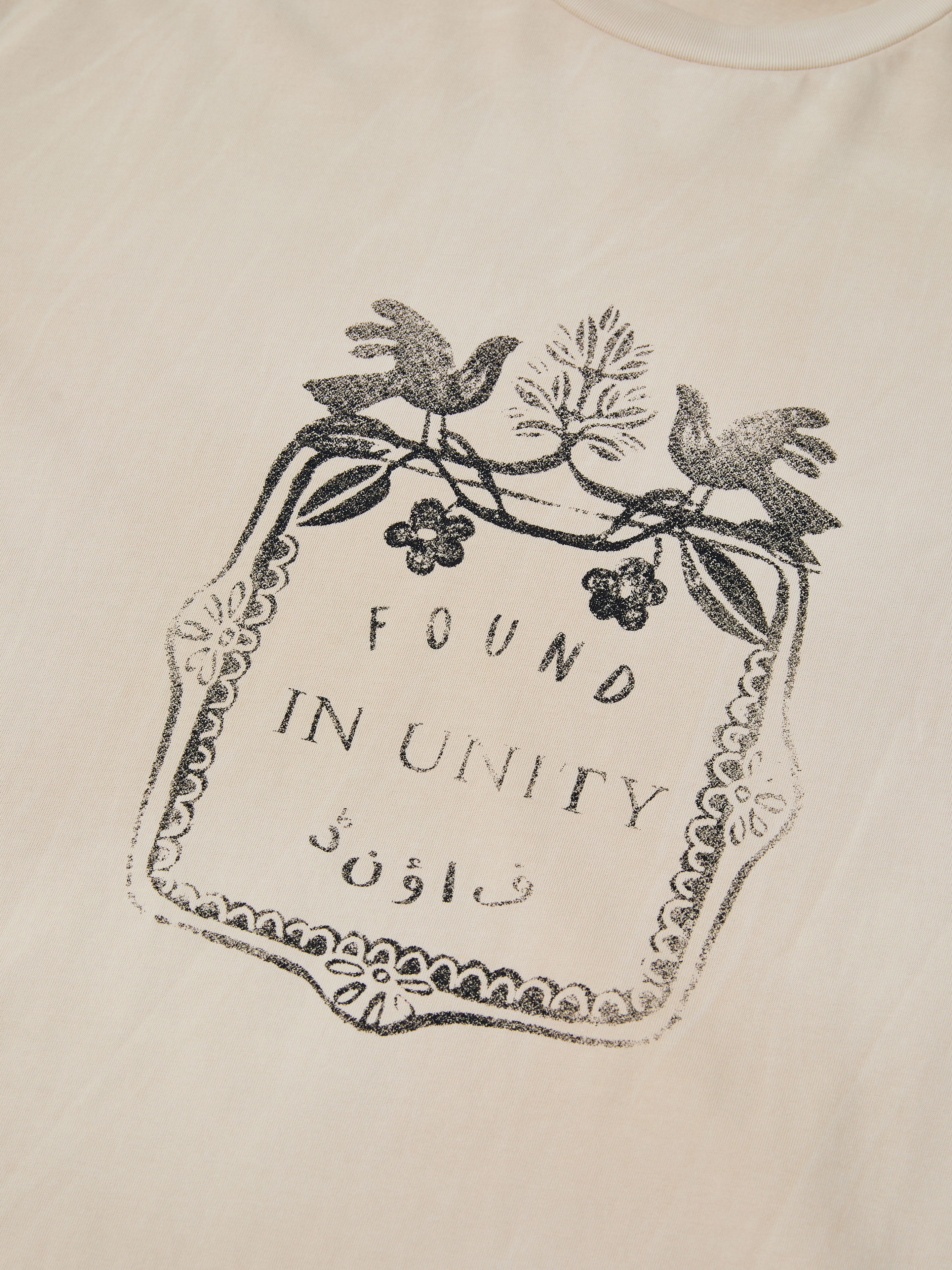 The UNITY CREST TEE by FOUND is a beige, mid-weight unisex cotton shirt with a black graphic—two birds, leaves, and “FOUND IN UNITY” in English and Urdu script inside a decorative border.