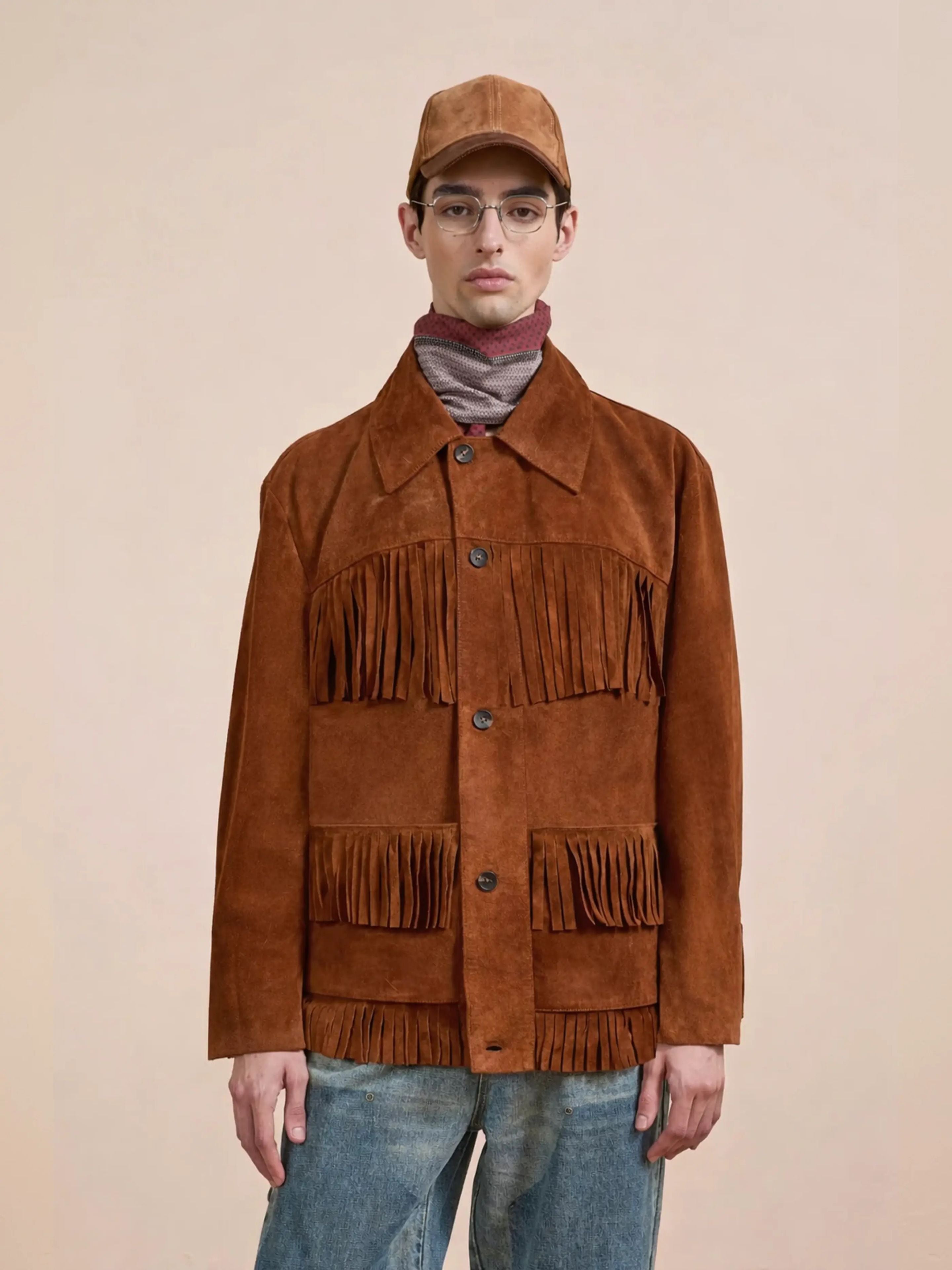 A person wears the FOUND Western Suede Fringe Jacket, a brown cap, striped scarf, glasses, and blue jeans while standing against a plain beige background.