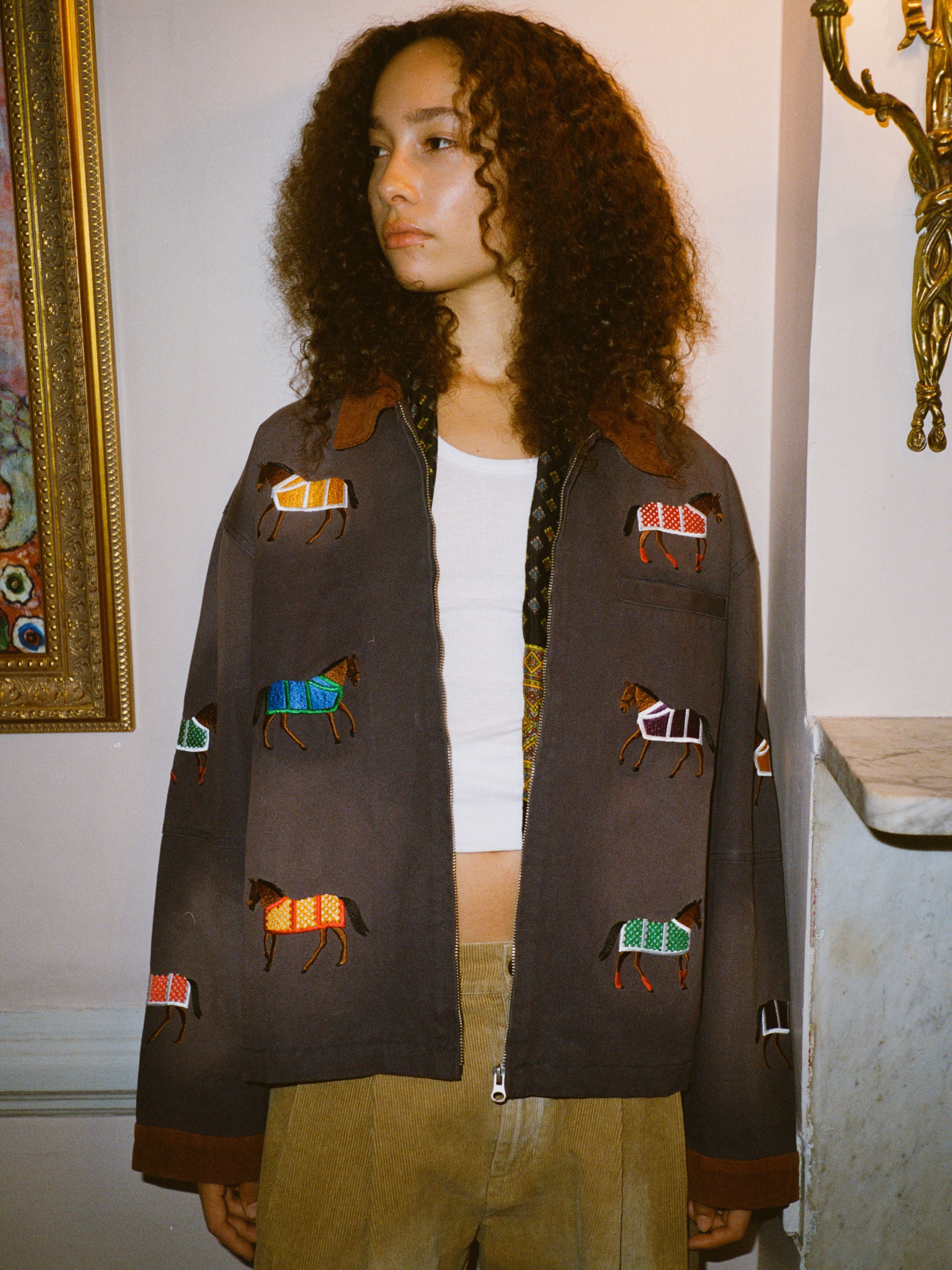 A person with curly hair wears the Found Lariat Horse Embroidered Work Jacket in navy over a white t-shirt, standing indoors beside a marble mantle and framed artwork.