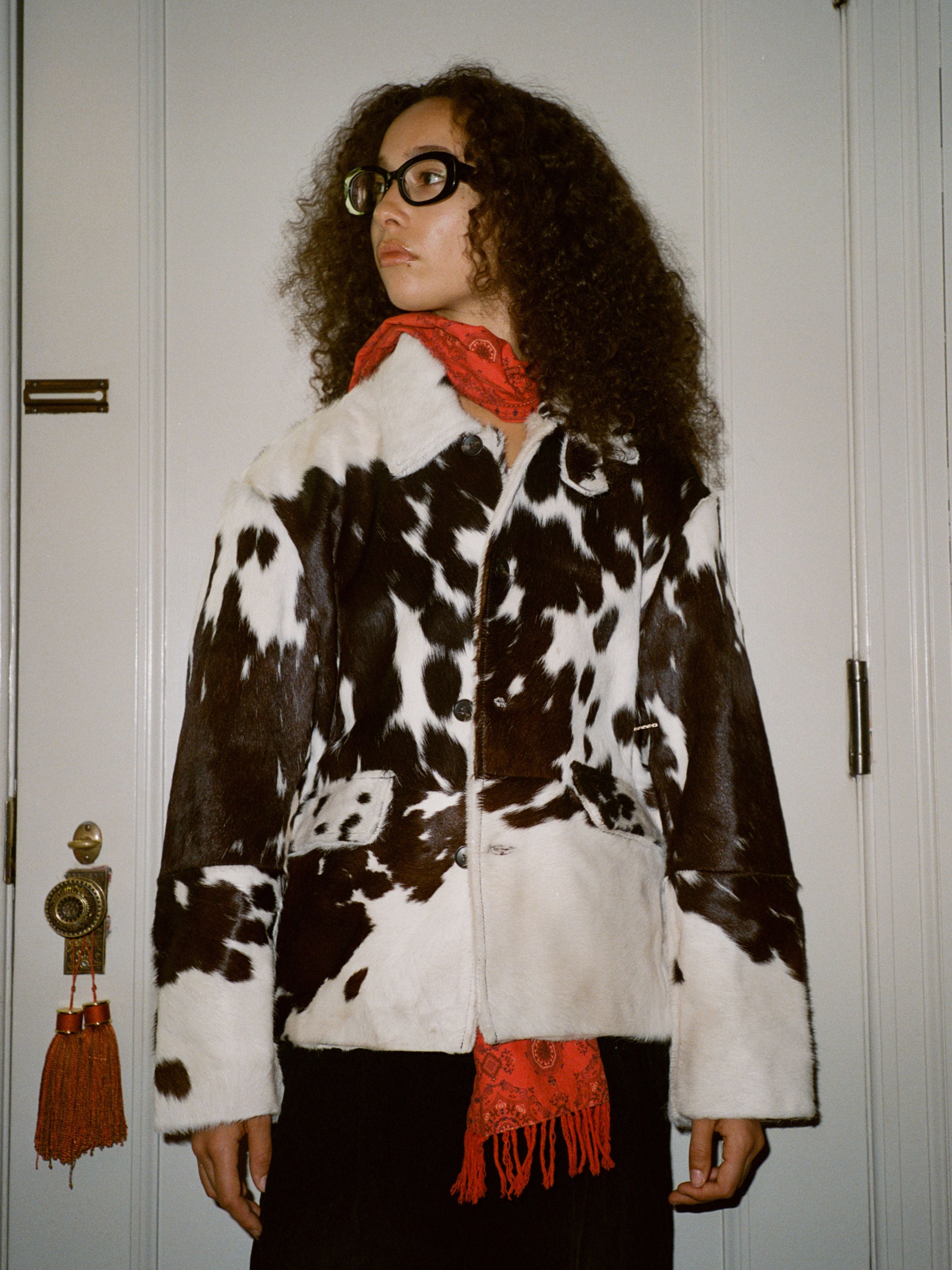 A person with curly hair and glasses wears the FOUND Cowhide Prairie Utility Jacket and a red scarf, standing before a white door with tassels on the handle.