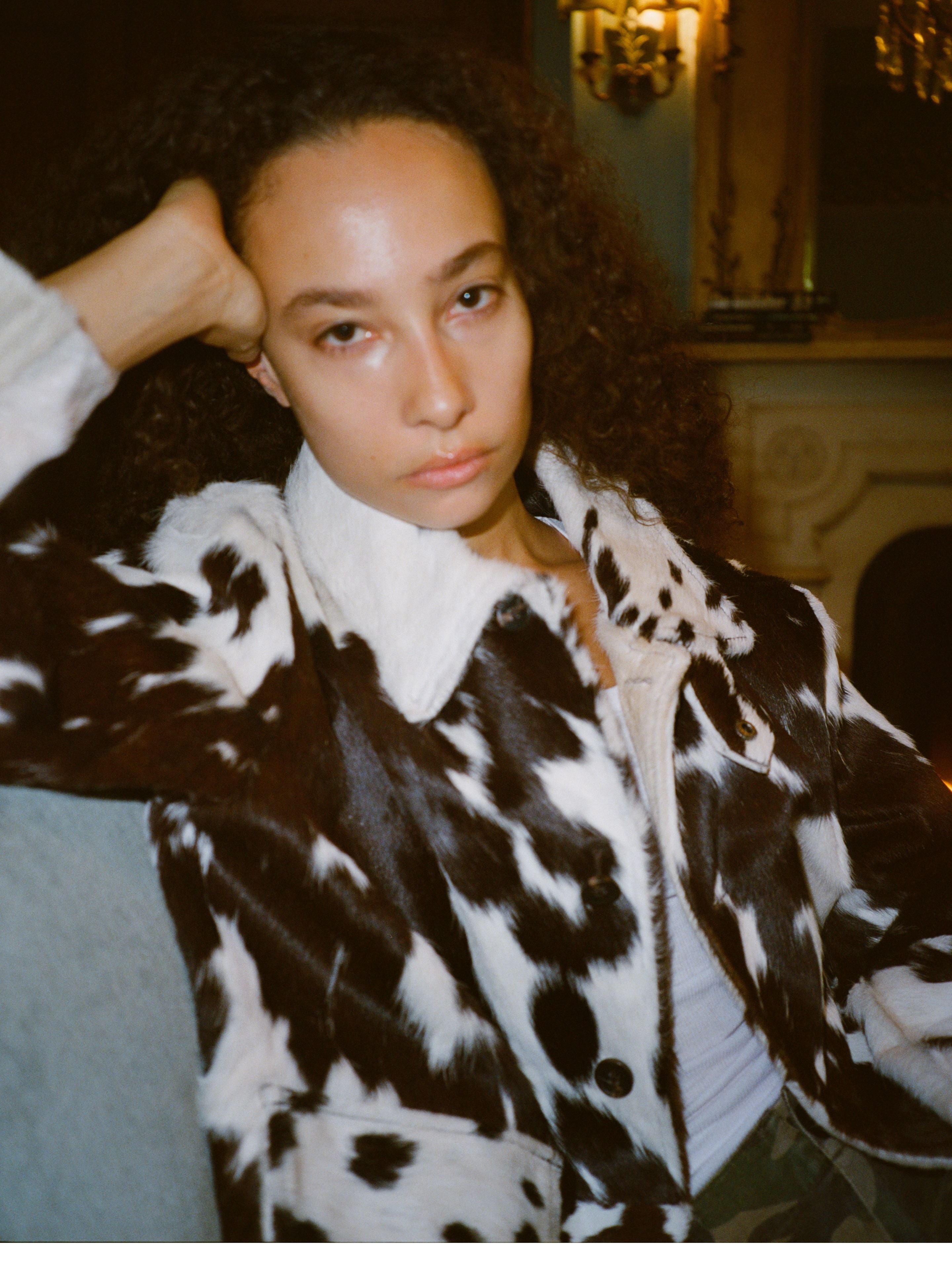 A person with curly hair wears the FOUND Cowhide Prairie Utility Jacket, sitting on a couch with their head resting on their hand in a dimly lit room.