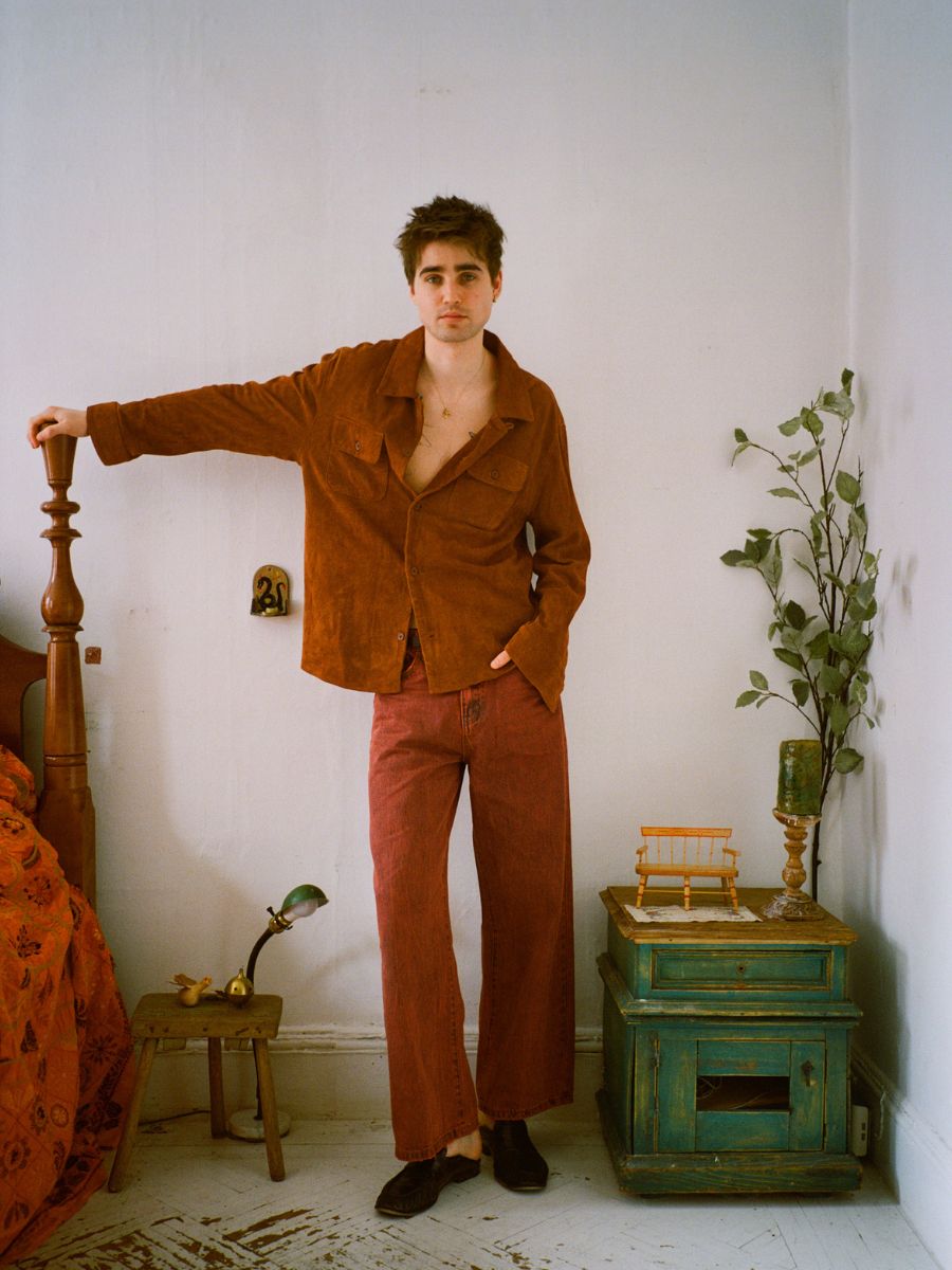 A man wearing the FOUND Suede Leather Camp Shirt in a unisex relaxed fit and wide red pants stands in a bedroom beside a bed, green nightstand, and potted plant against a white wall.