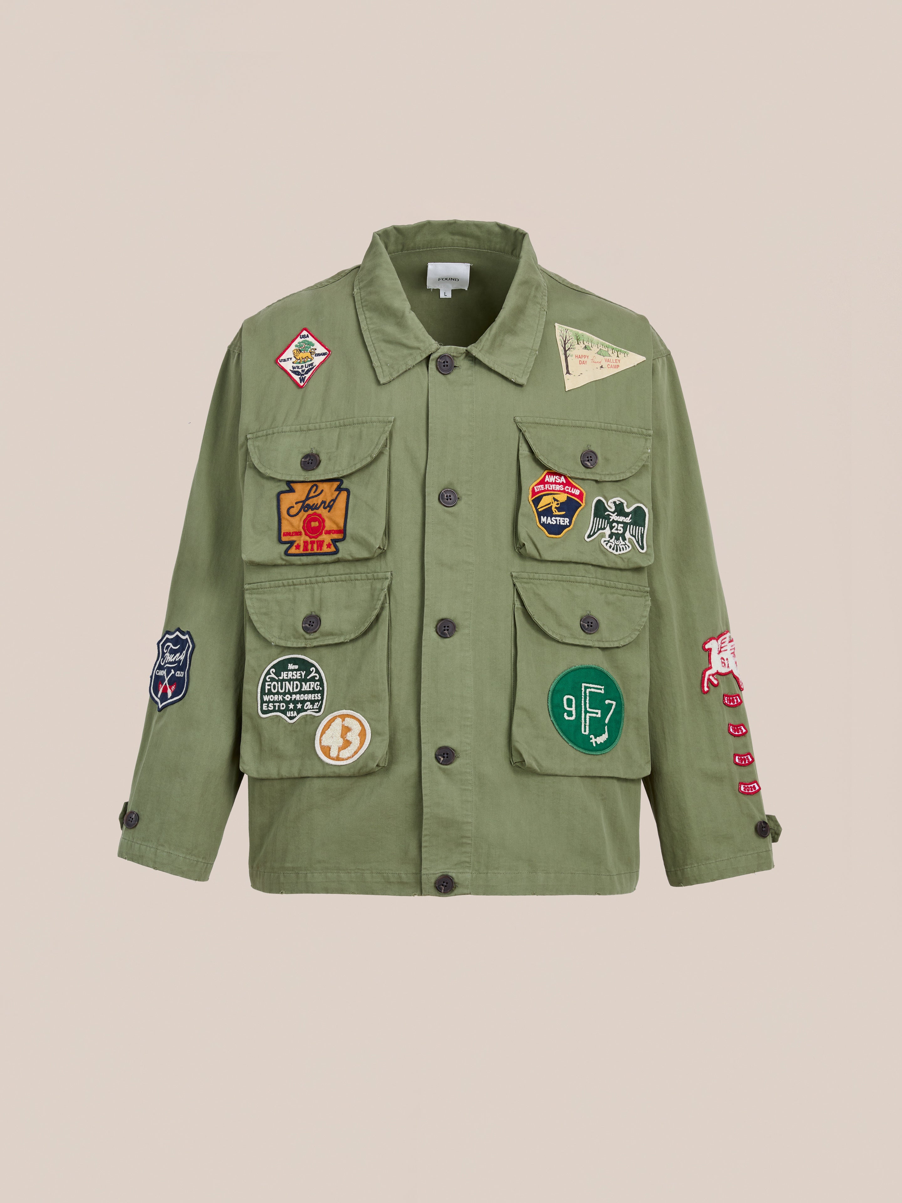 The FOUND Herringbone Utility Patchwork Jacket is a military-inspired olive green cotton jacket with multiple front pockets and colorful embroidered patches, shown against a plain beige background.