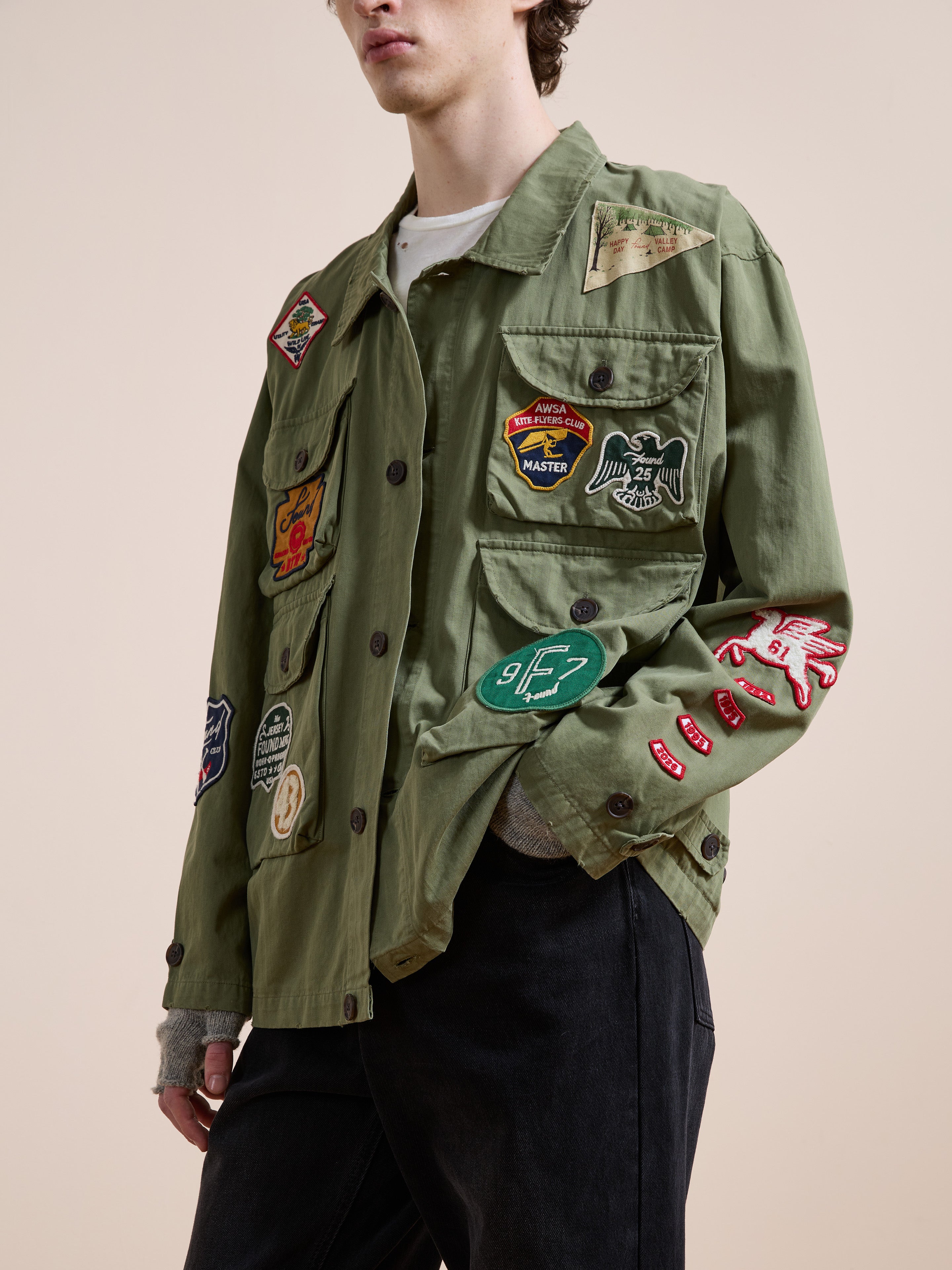 A person wears the FOUND Herringbone Utility Patchwork Jacket, featuring colorful patches, over a white shirt and dark pants, standing against a plain beige background.