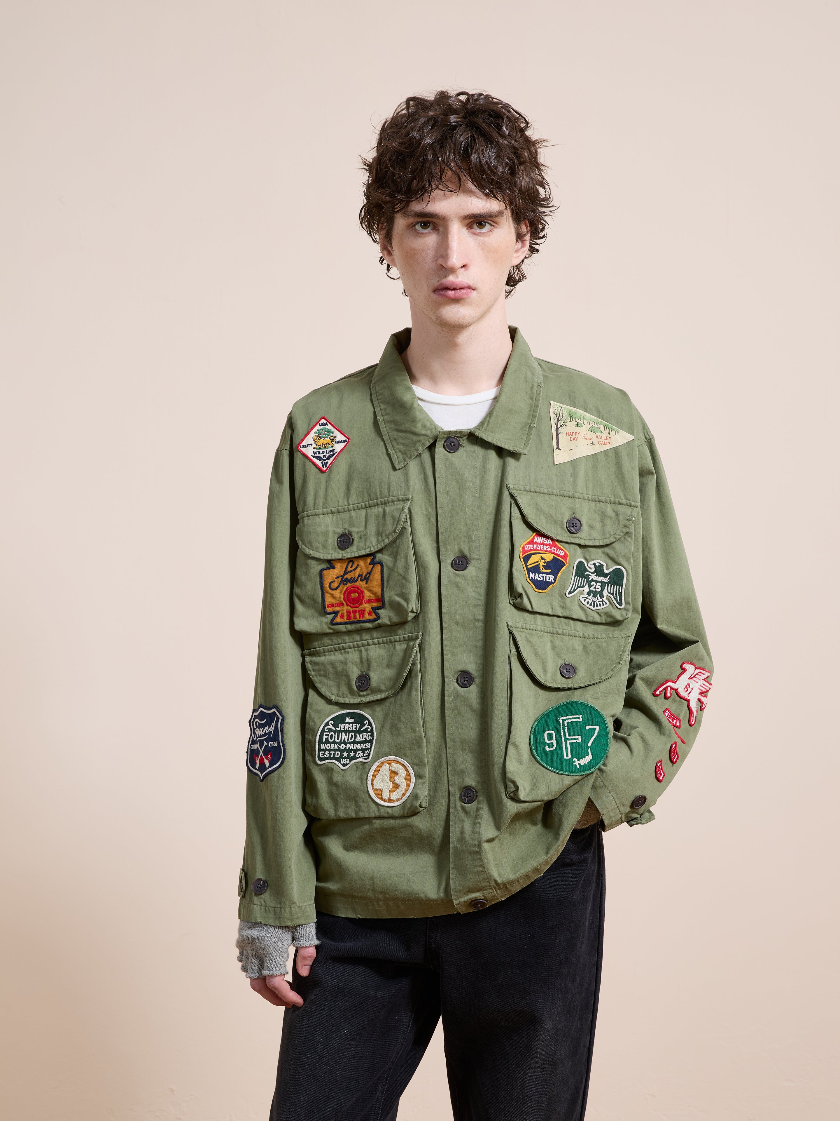 A person with short curly hair wears the FOUND Herringbone Utility Patchwork Jacket in olive green, featuring colorful patches, and stands against a plain beige background.