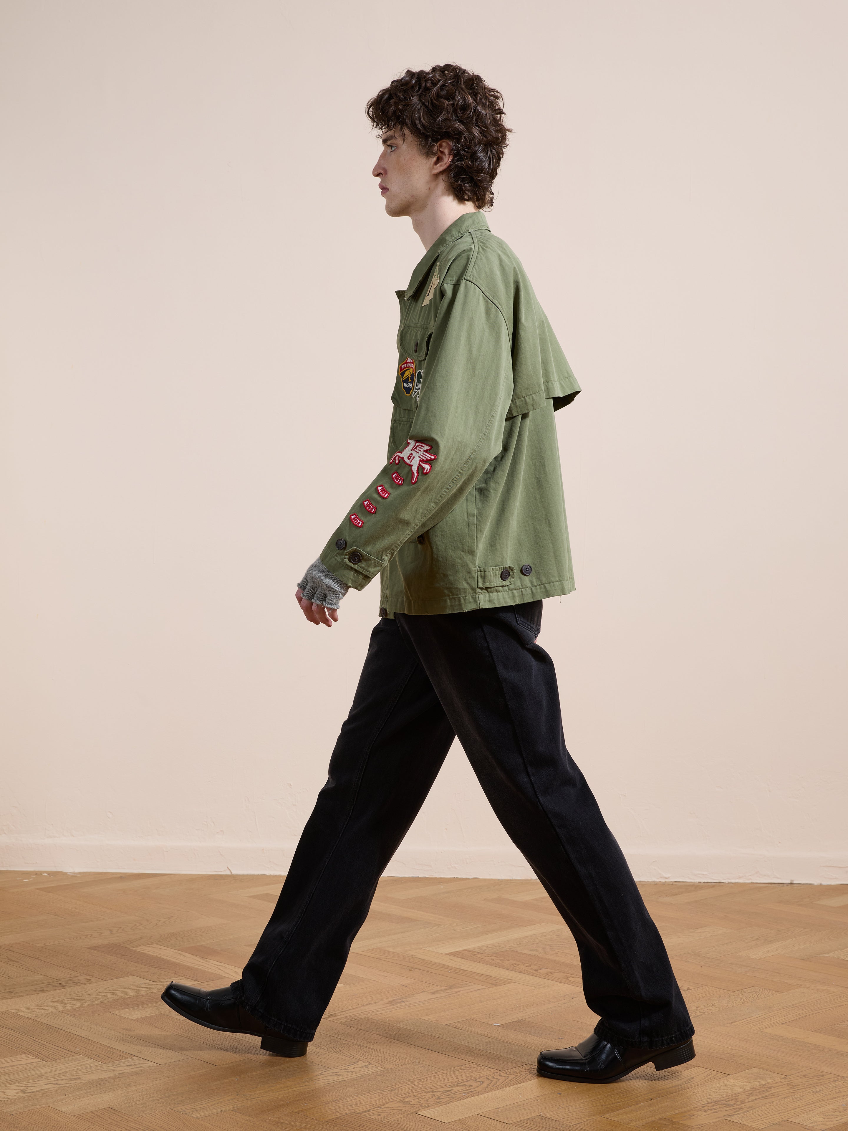 A person with curly hair wears the FOUND Herringbone Utility Patchwork Jacket, paired with black pants and shoes, standing on a wooden floor against a plain beige wall.
