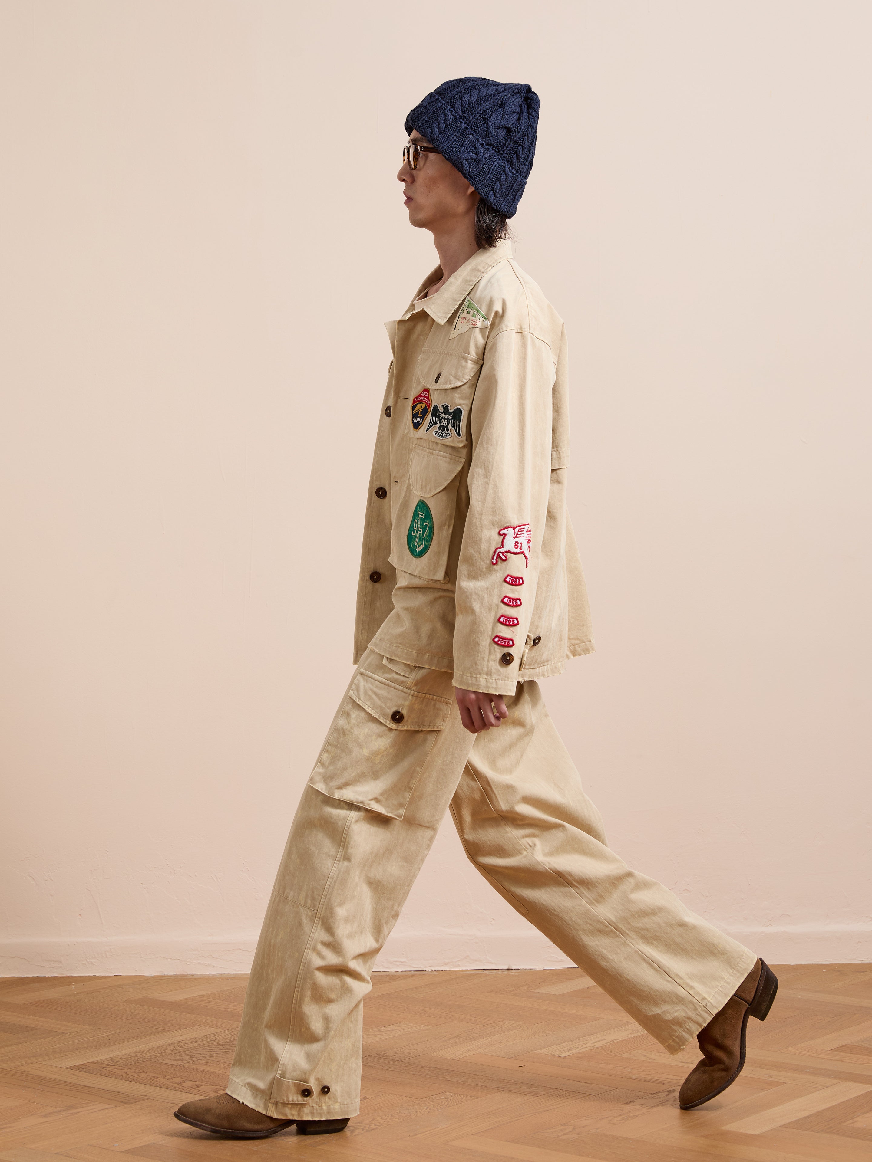 A person in the FOUND Herringbone Utility Patchwork Jacket, a navy knit hat, brown boots, and glasses walks indoors on a wooden floor.