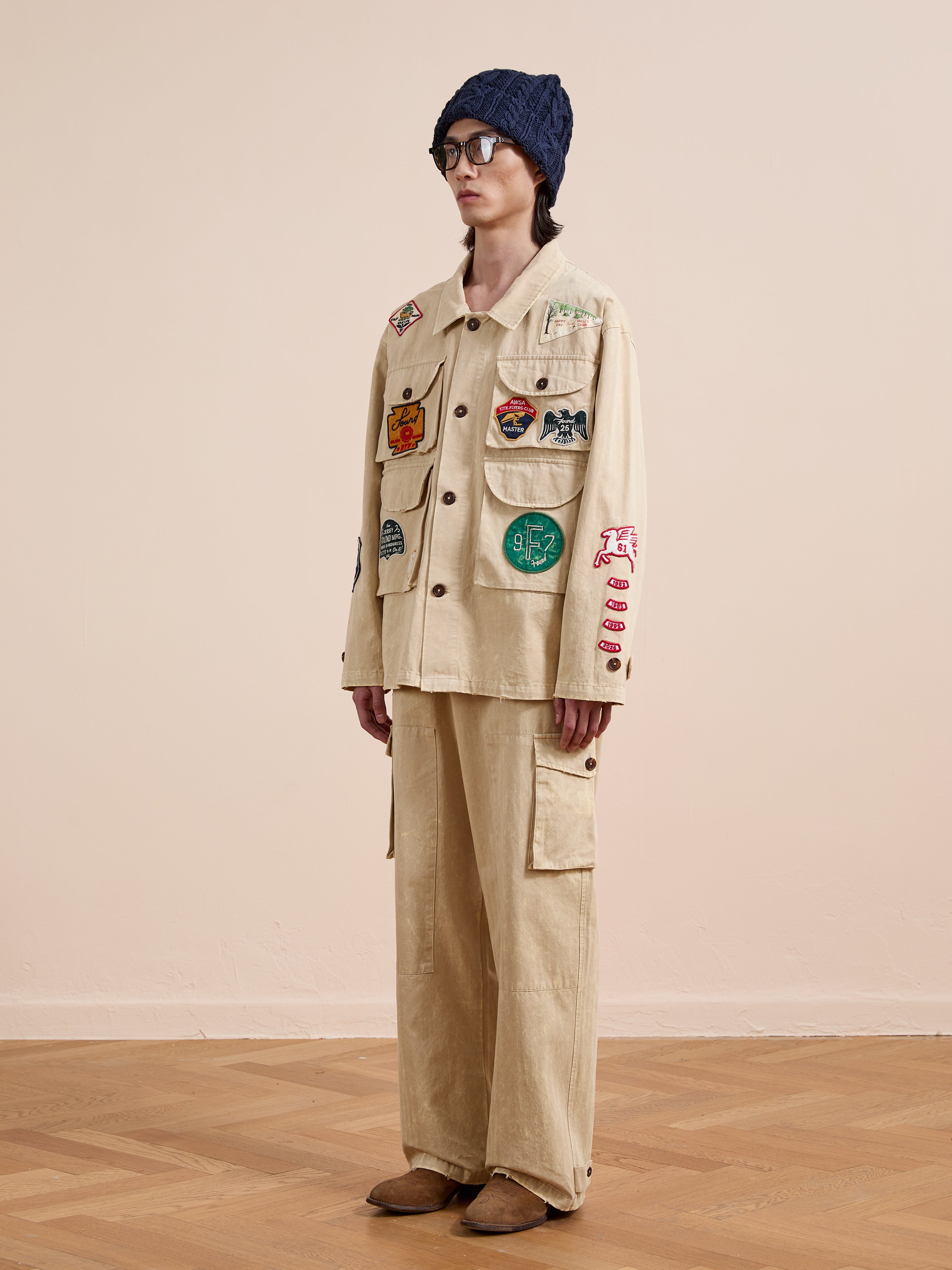 Indoors against a plain wall and wooden floor, a person wears FOUND’s Herringbone Utility Patchwork Jacket with colorful patches, matching pants, a navy knit hat, glasses, and brown shoes.