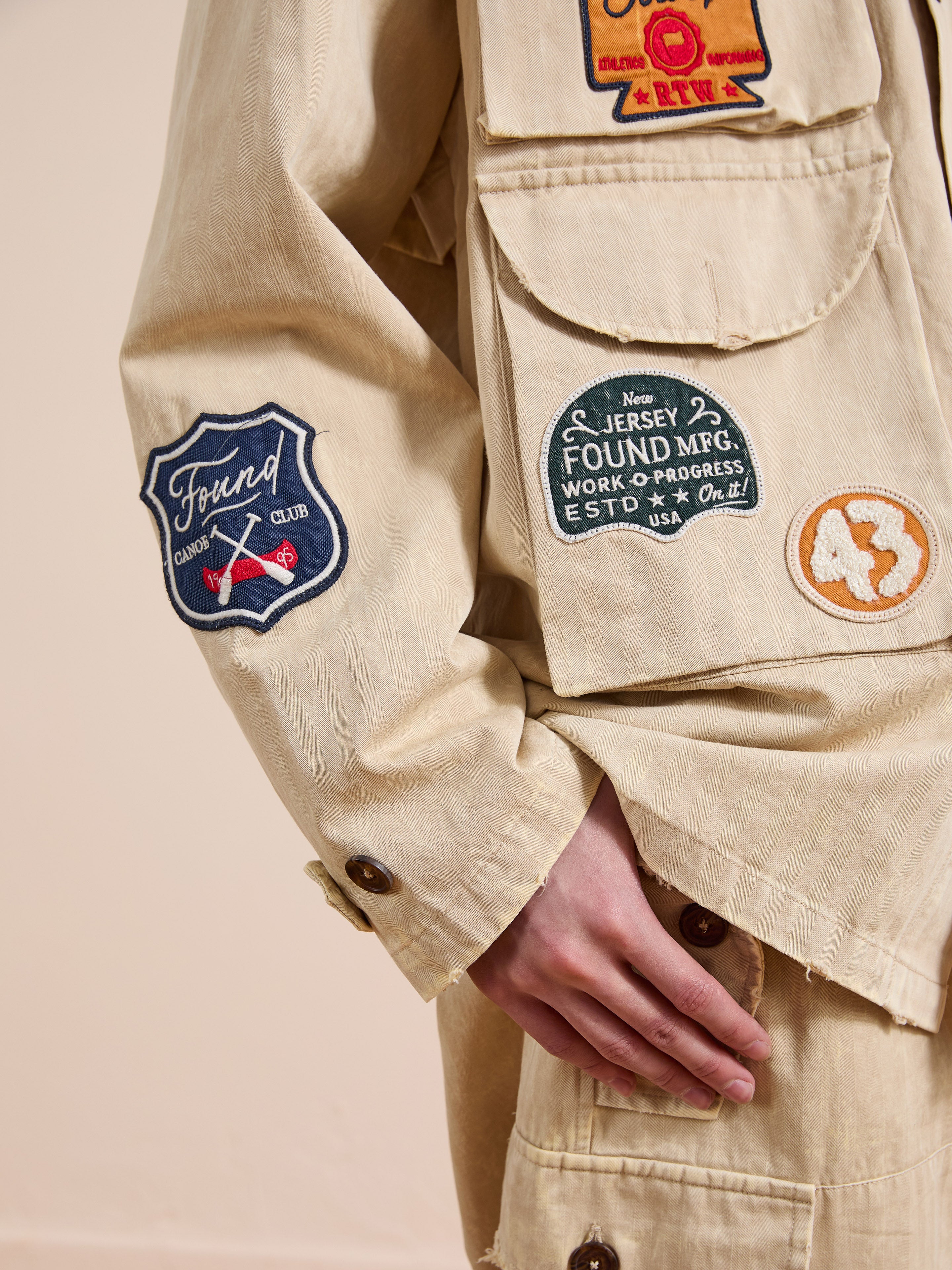 A person wears the FOUND Herringbone Utility Patchwork Jacket in beige, featuring multiple colorful embroidered patches, badges, and numbers, with one hand resting by their side.