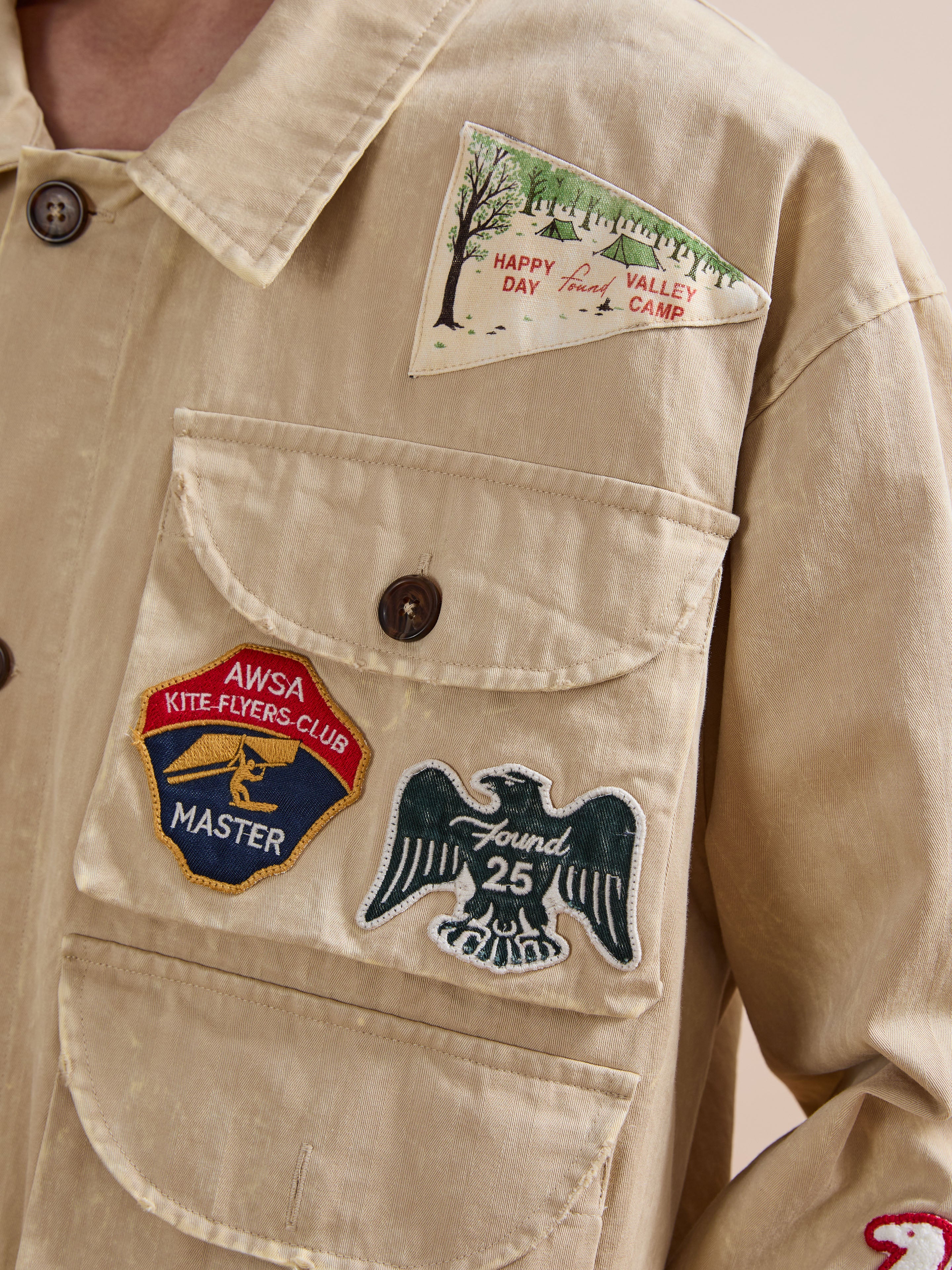 The FOUND Herringbone Utility Patchwork Jacket is a unisex vintage-inspired canvas jacket, adorned with embroidered patches like an eagle, a kite-flyers club badge, and a "Happy Day Valley Camp" pennant—blending utility style and playful charm.
