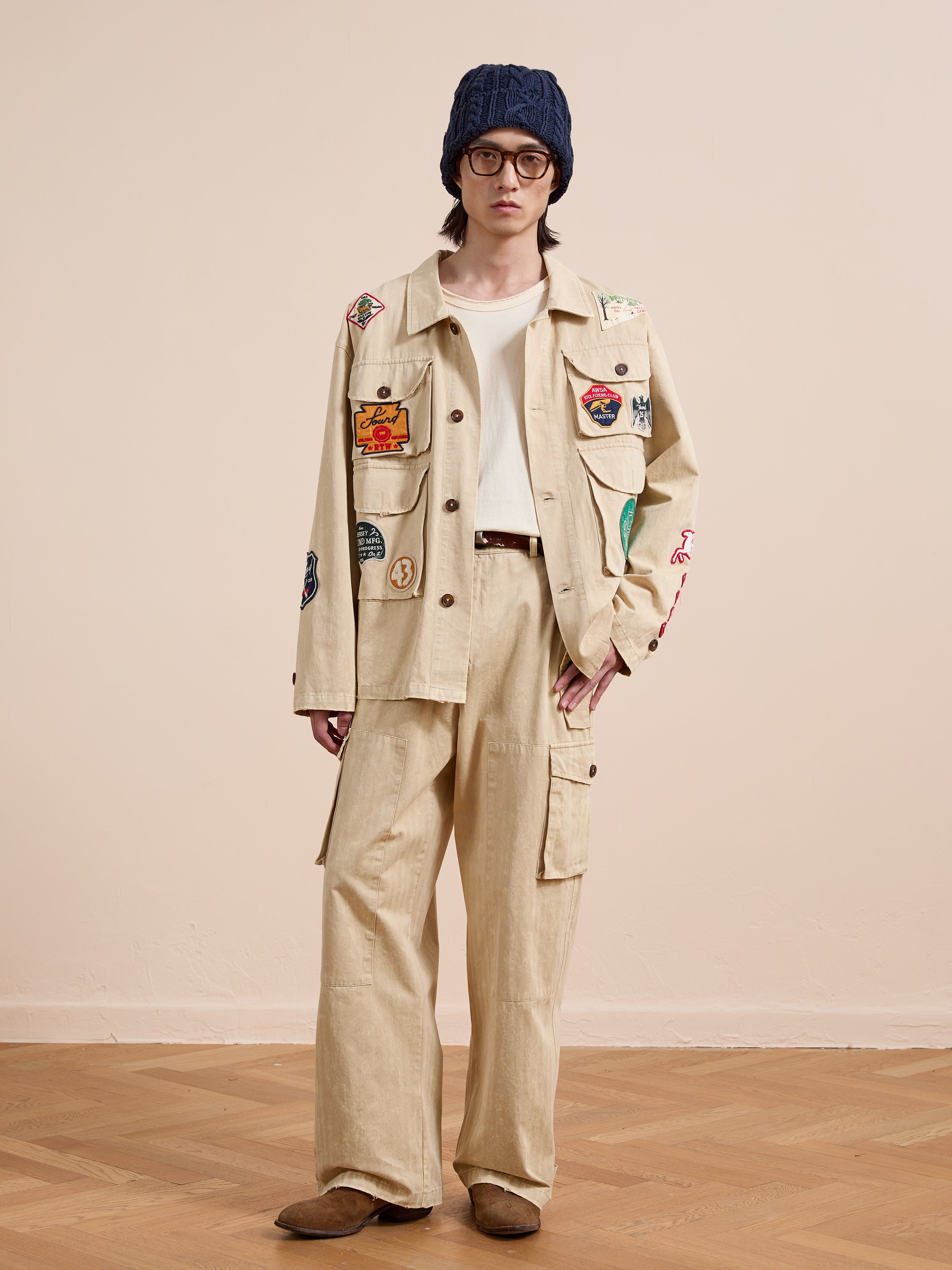 A person stands indoors on a wooden floor against a plain wall, wearing FOUND's Herringbone Utility Patchwork Jacket, matching cargo pants, white shirt, blue knit beanie, round glasses, and brown shoes.