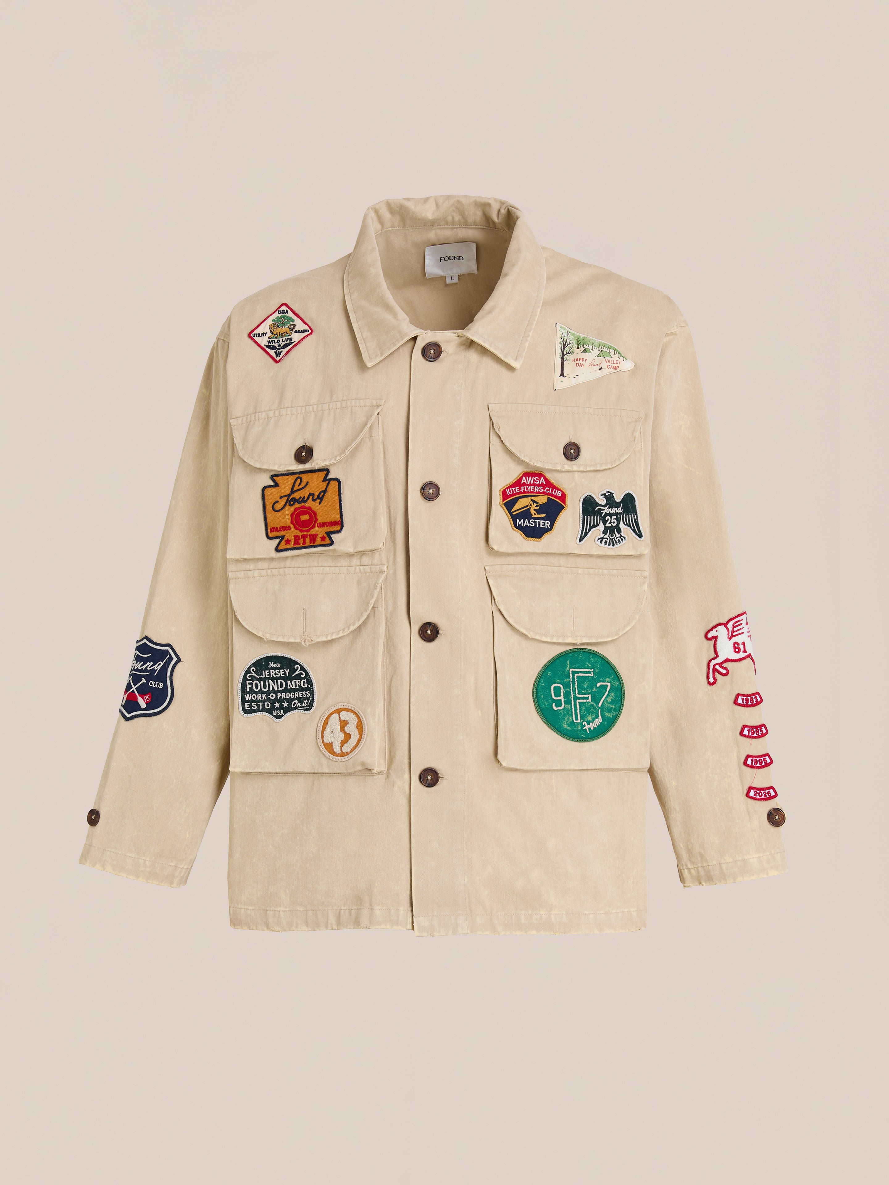 FOUND Herringbone Utility Patchwork Jacket in beige canvas features brown buttons, four front pockets, and colorful embroidered patches on the front, sleeves, and pockets, shown on a plain background.