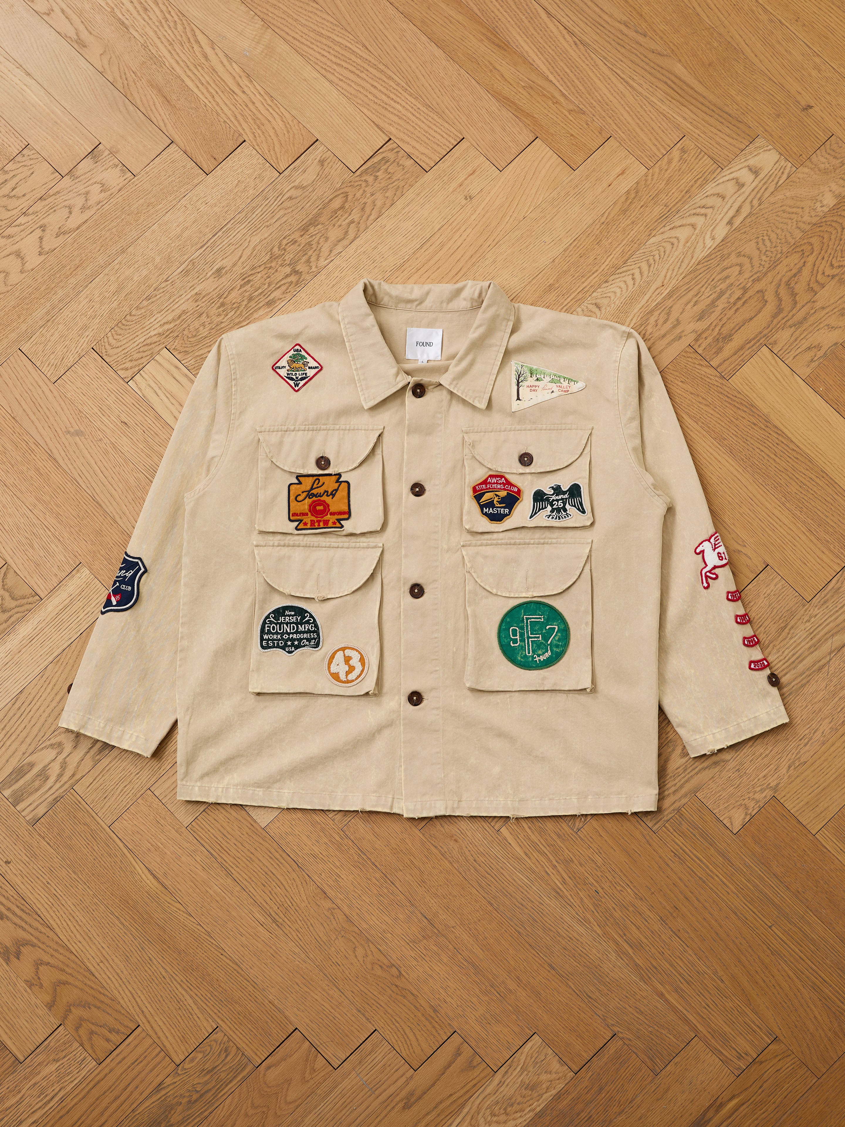 The FOUND Herringbone Utility Patchwork Jacket is a unisex vintage beige canvas piece featuring colorful embroidered patches on the front, sleeves, and pockets, displayed flat on a wooden herringbone floor.