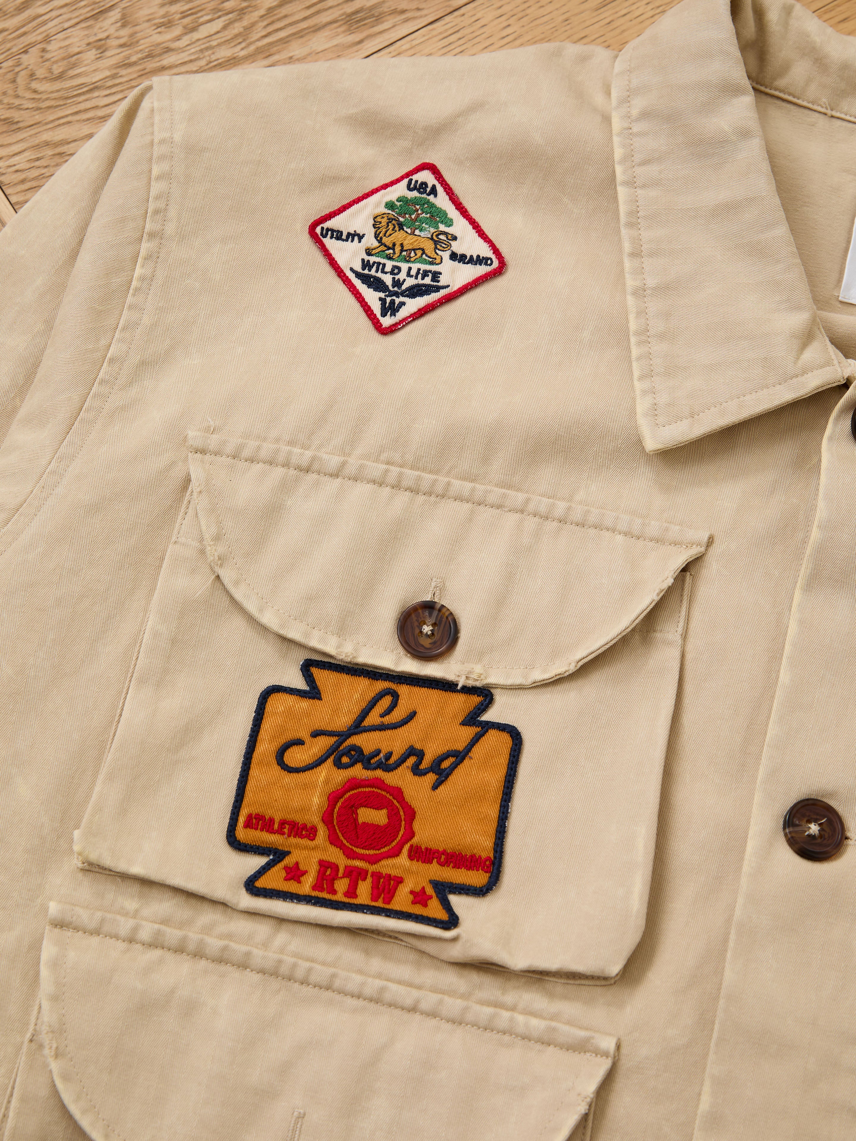 The FOUND Herringbone Utility Patchwork Jacket is a beige shirt-style jacket with two buttoned chest pockets and embroidered animal and brand patches on the left side.
