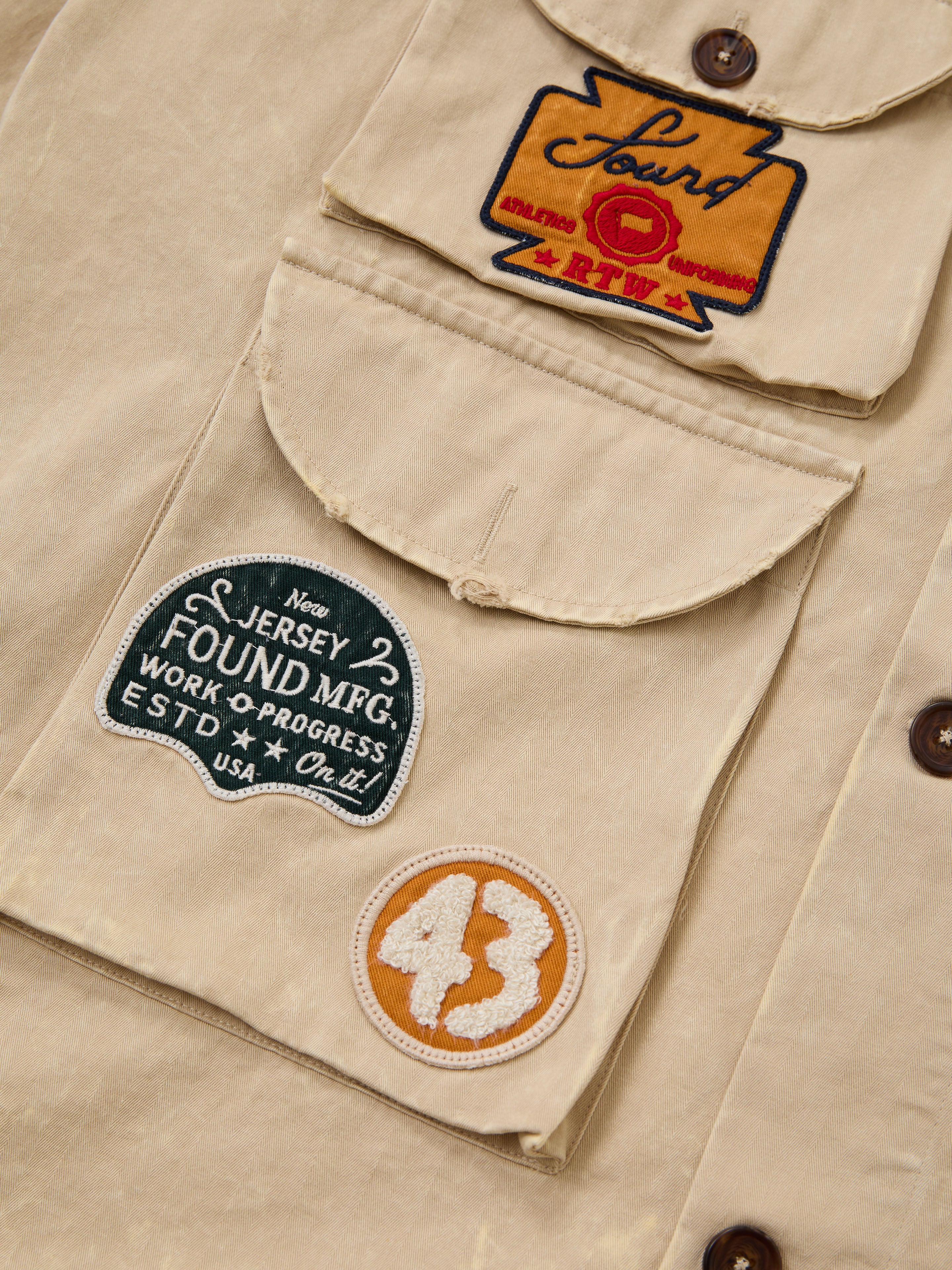 Close-up of the FOUND Herringbone Utility Patchwork Jacket, featuring three stitched patches: "Lourd RTW," "New Jersey Found MFG. ESTD USA," and a circular orange patch with the number 43.
