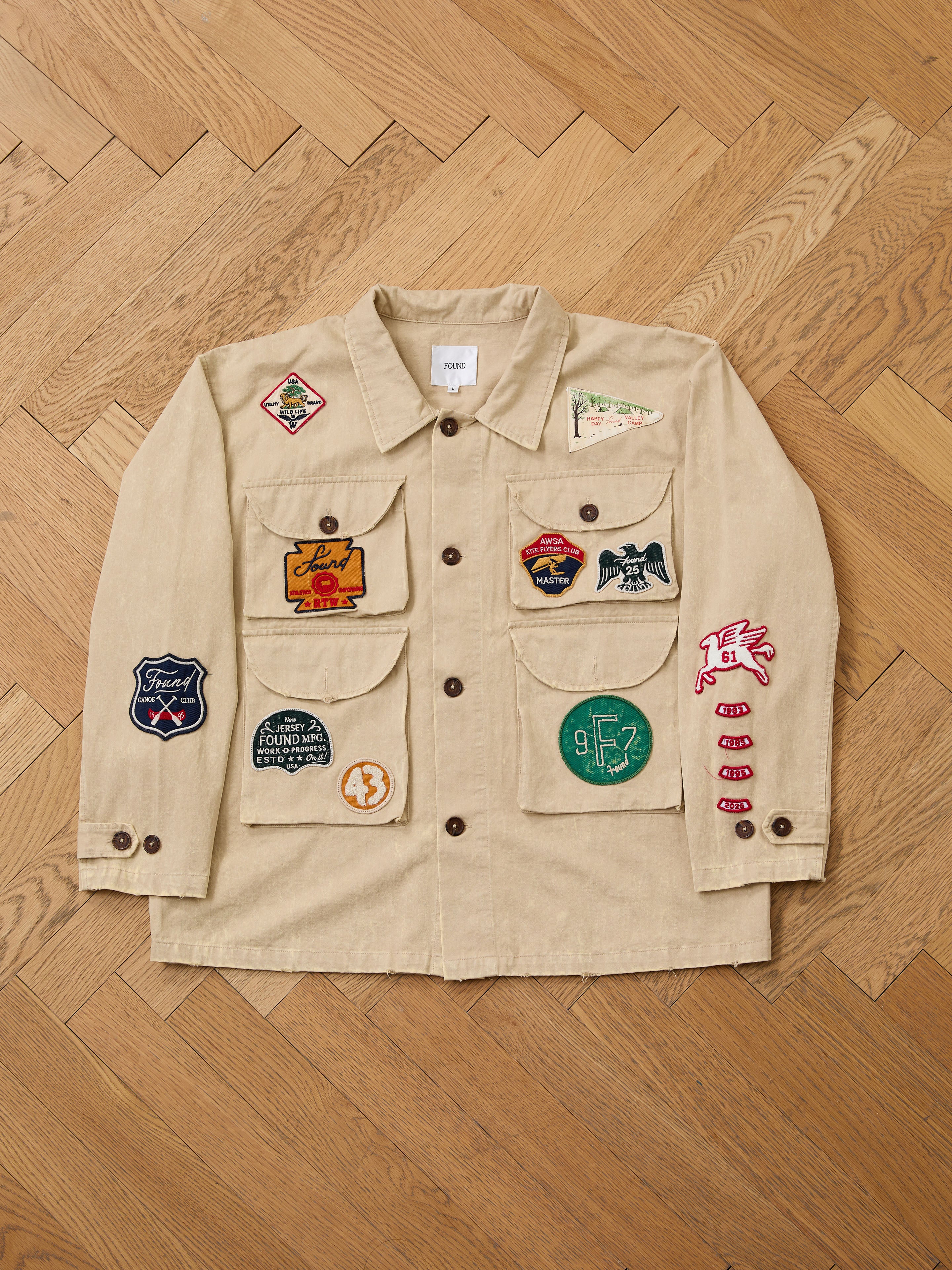The FOUND Herringbone Utility Patchwork Jacket—a beige vintage canvas piece with colorful embroidered patches—offers unique, unisex style. Displayed on a wooden herringbone floor, it blends utility wear with standout appeal.
