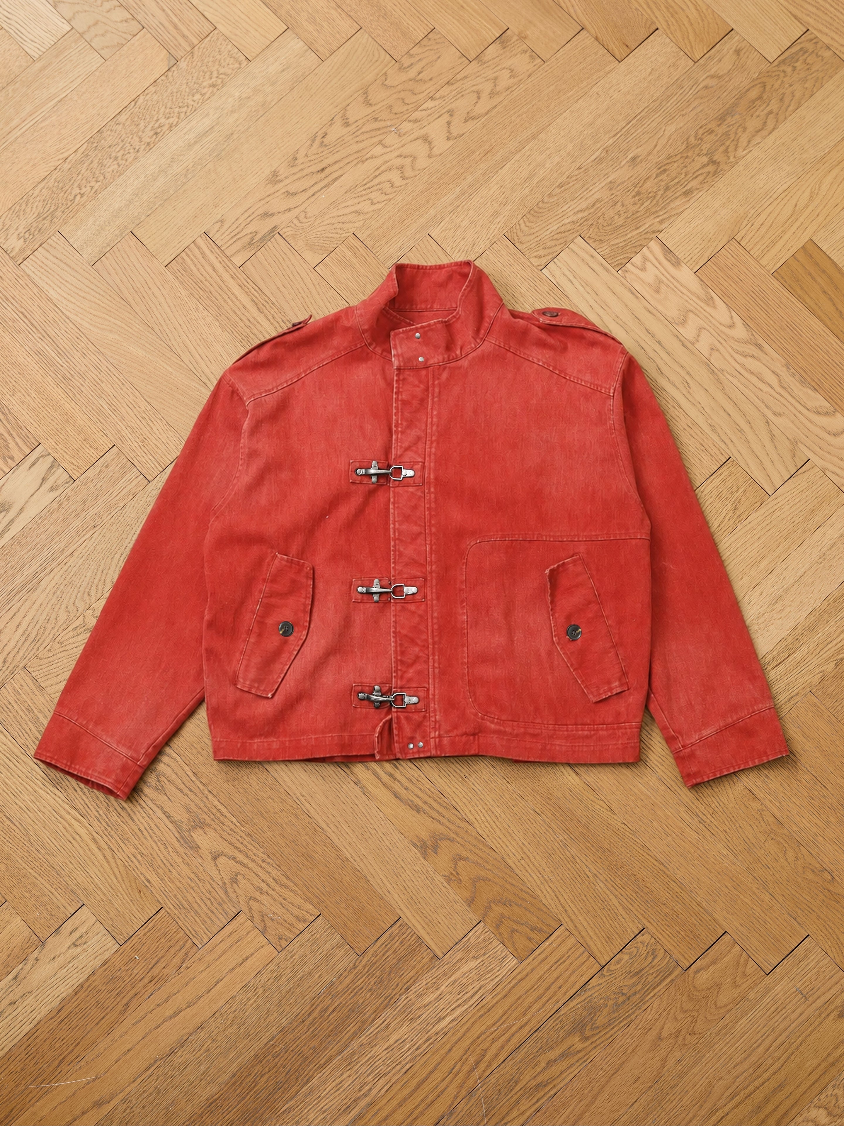 The FOUND Utility Clasp Work Jacket, a vintage-inspired red piece featuring long sleeves, front toggle closures reminiscent of fireman clasps, two flap pockets, and distinctive button details, is laid flat on a herringbone-patterned wooden floor.