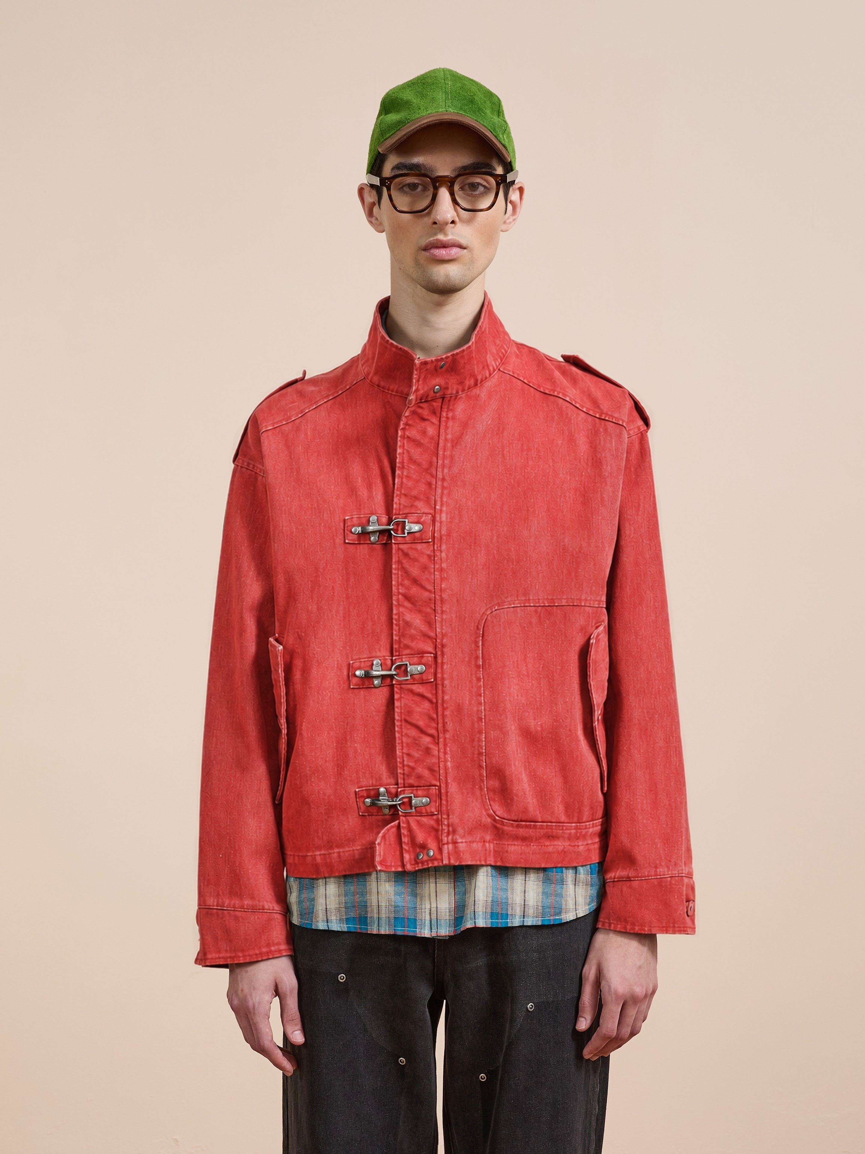 A person wearing the FOUND Utility Clasp Work Jacket in vintage red, paired with a green cap, glasses, plaid shirt, and dark pants stands against a plain light background.