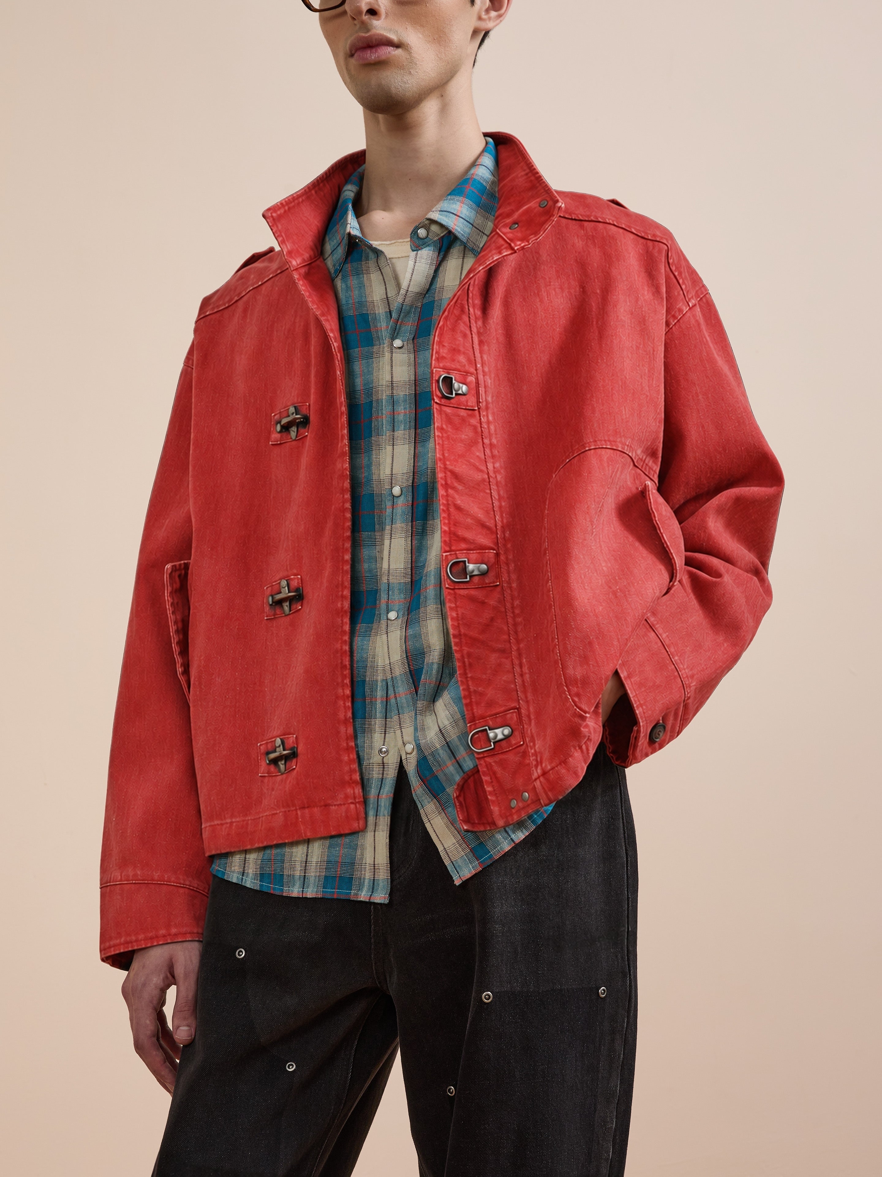 Model wearing the FOUND Utility Clasp Work Jacket—a vintage-inspired red jacket with metal fireman clasps—layered over a blue plaid shirt and black pants, standing against a plain beige background.
