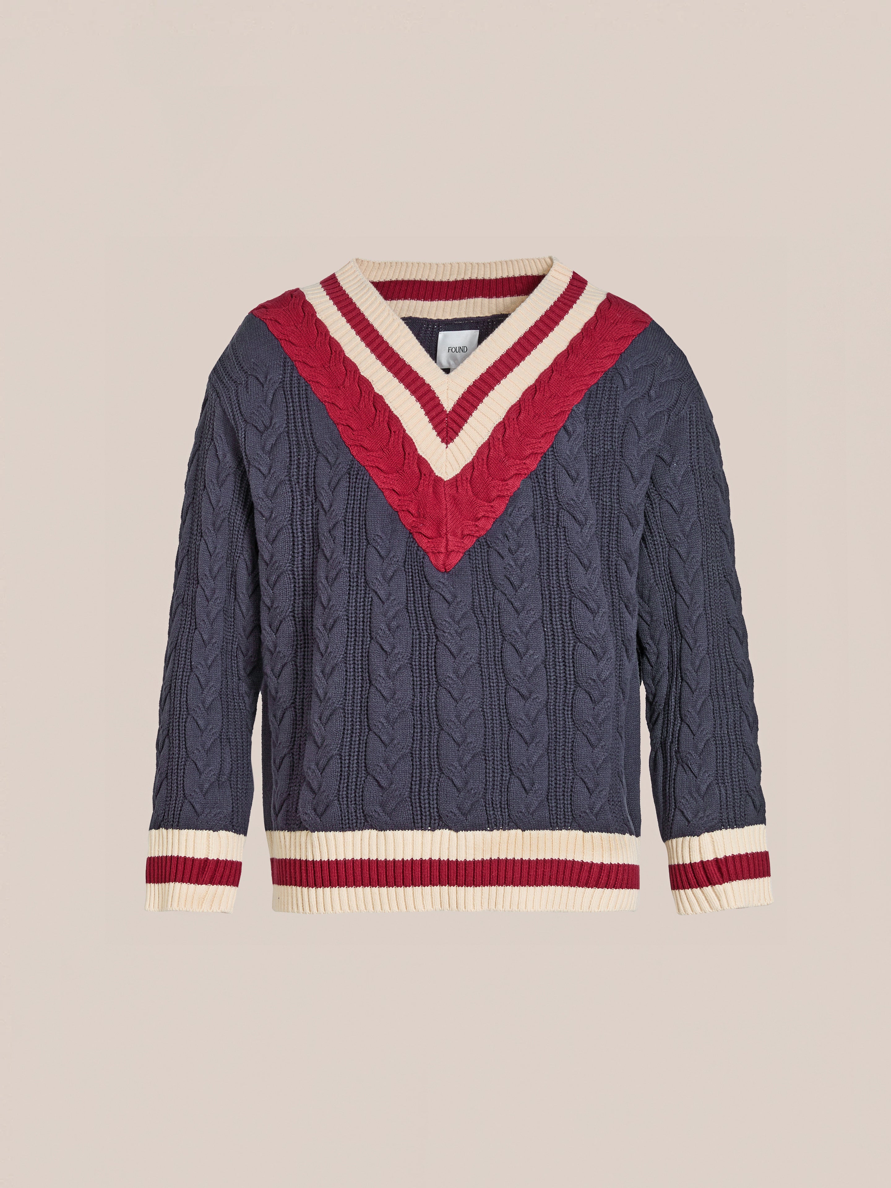 The FOUND V-Neck Cricket Sweater is navy with a cable-knit design, featuring a red and cream chevron pattern on the chest. It has matching sleeve trim and is crafted from soft cotton for ultimate comfort.