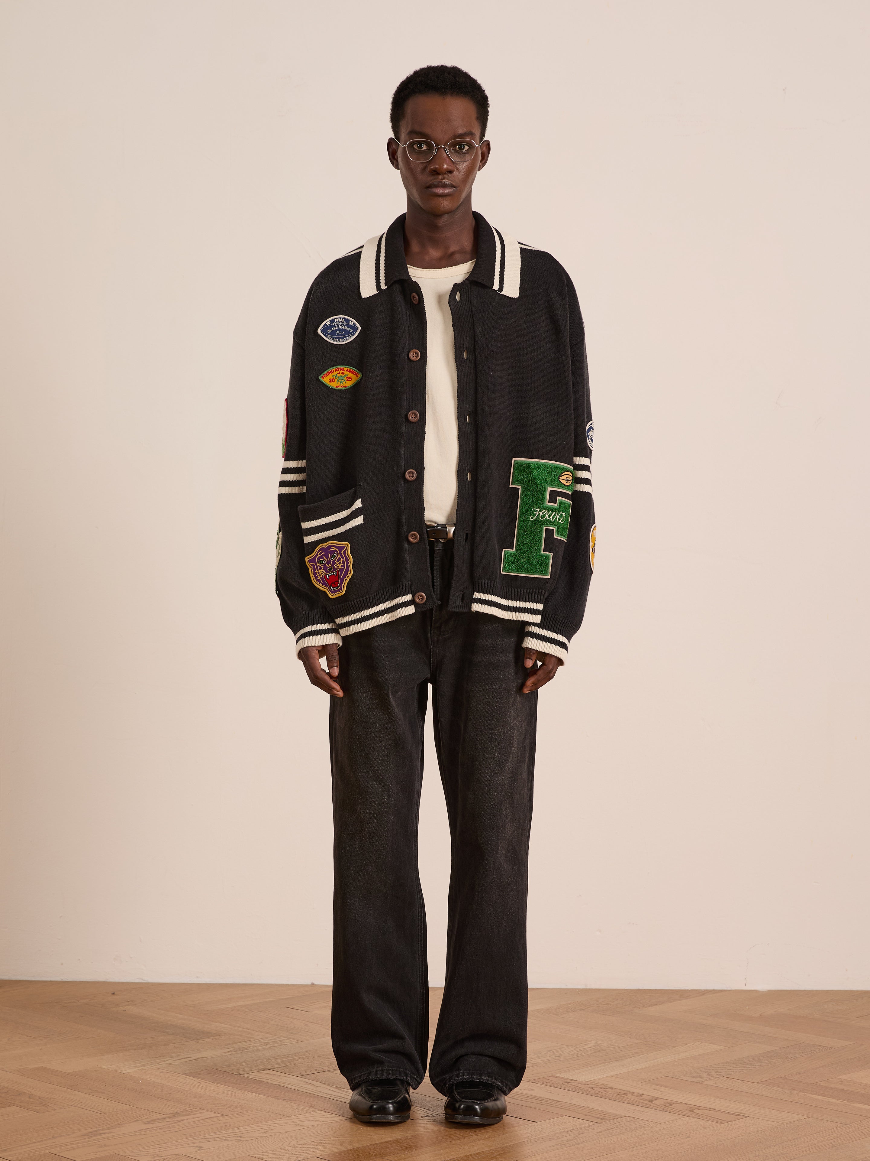 A person stands indoors against a plain light background, wearing FOUND's Varsity Patch Collared Cardigan over a white shirt, paired with black wide-leg jeans, round glasses, and black shoes.