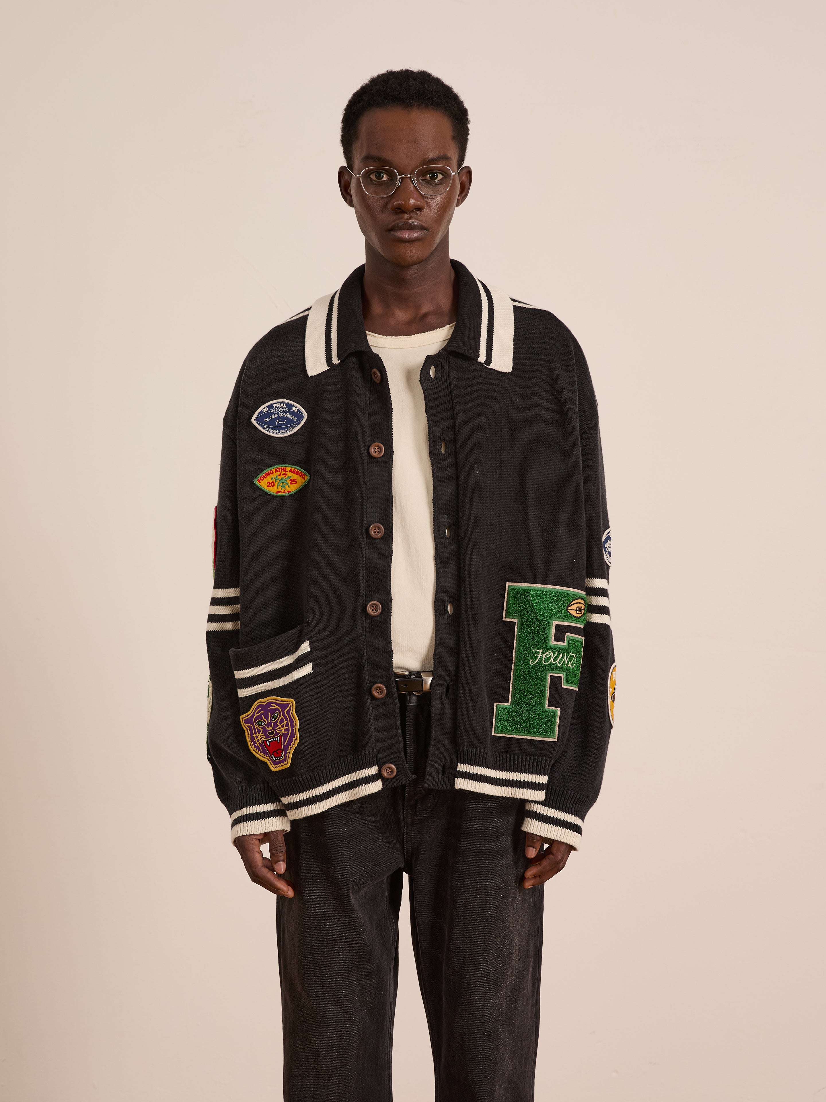 A person wearing glasses, a white shirt, and the FOUND Varsity Patch Collared Cardigan stands against a plain light-colored background.