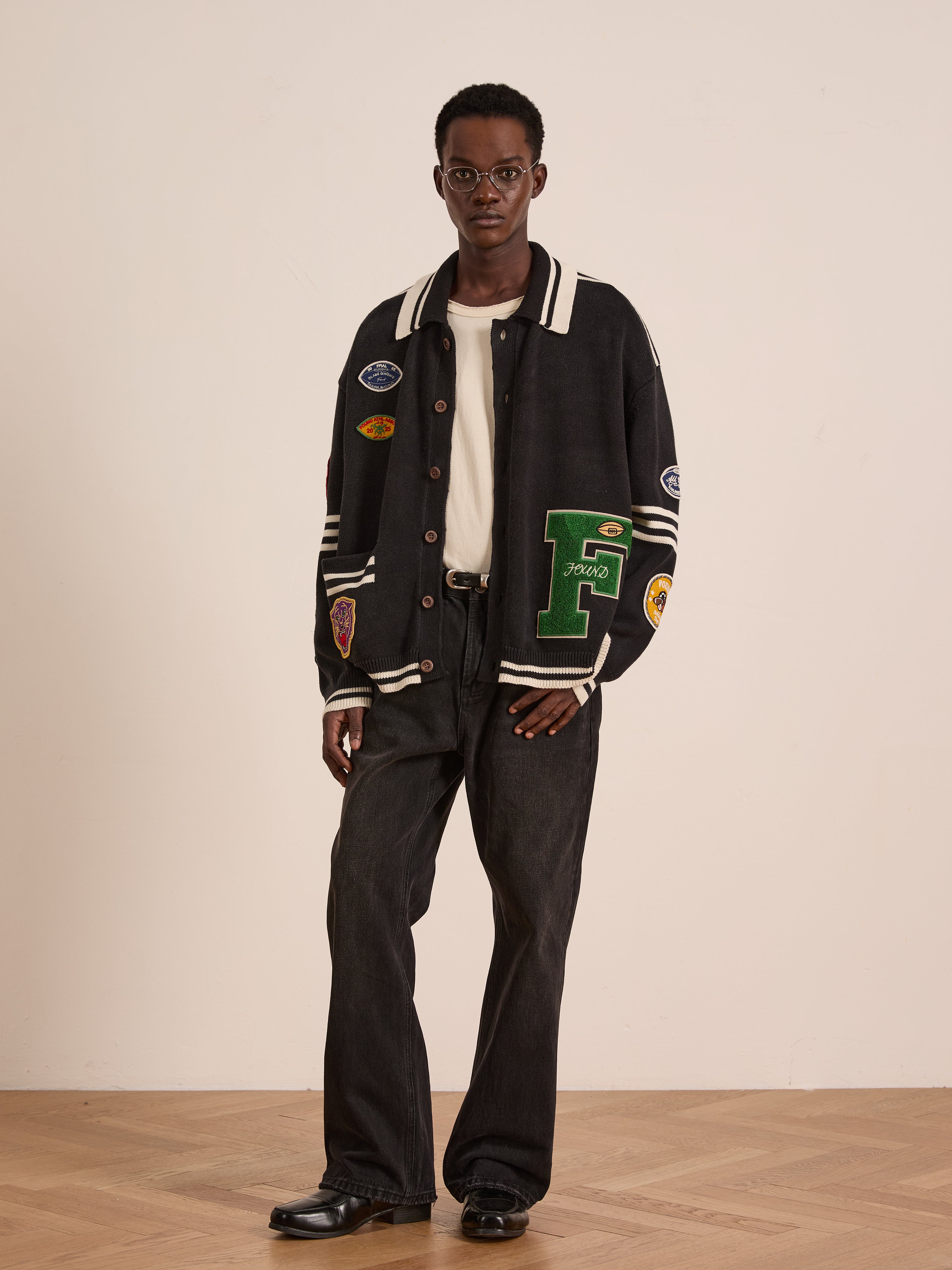 A man stands indoors wearing glasses, a white shirt, black jeans, black shoes, and the FOUND Varsity Patch Collared Cardigan. He faces forward with a neutral expression, highlighting the cardigan’s collegiate-inspired style.