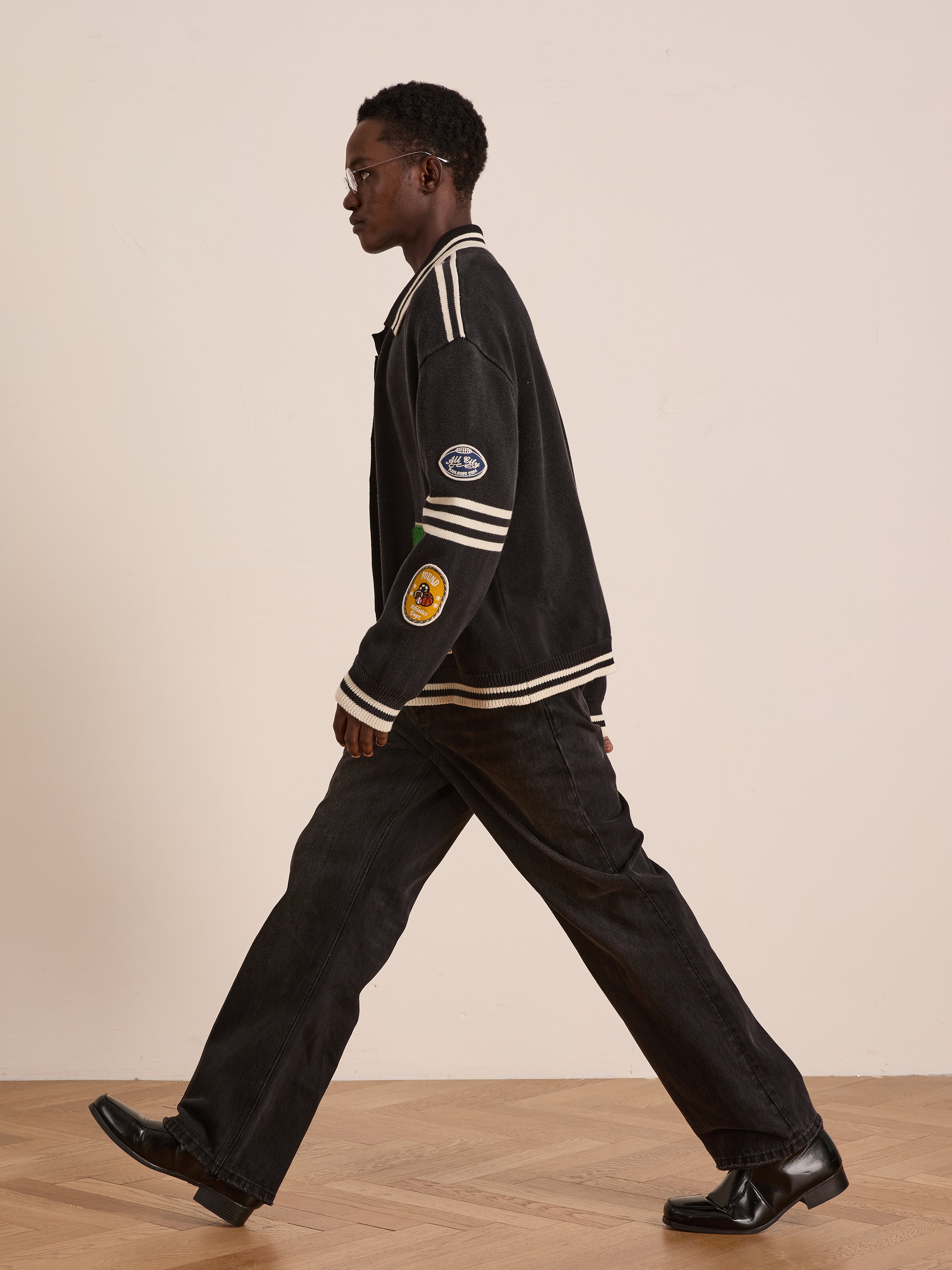 A person wearing dark clothing, black shoes, and the FOUND Varsity Patch Collared Cardigan walks indoors on a wooden floor against a plain light-colored wall.