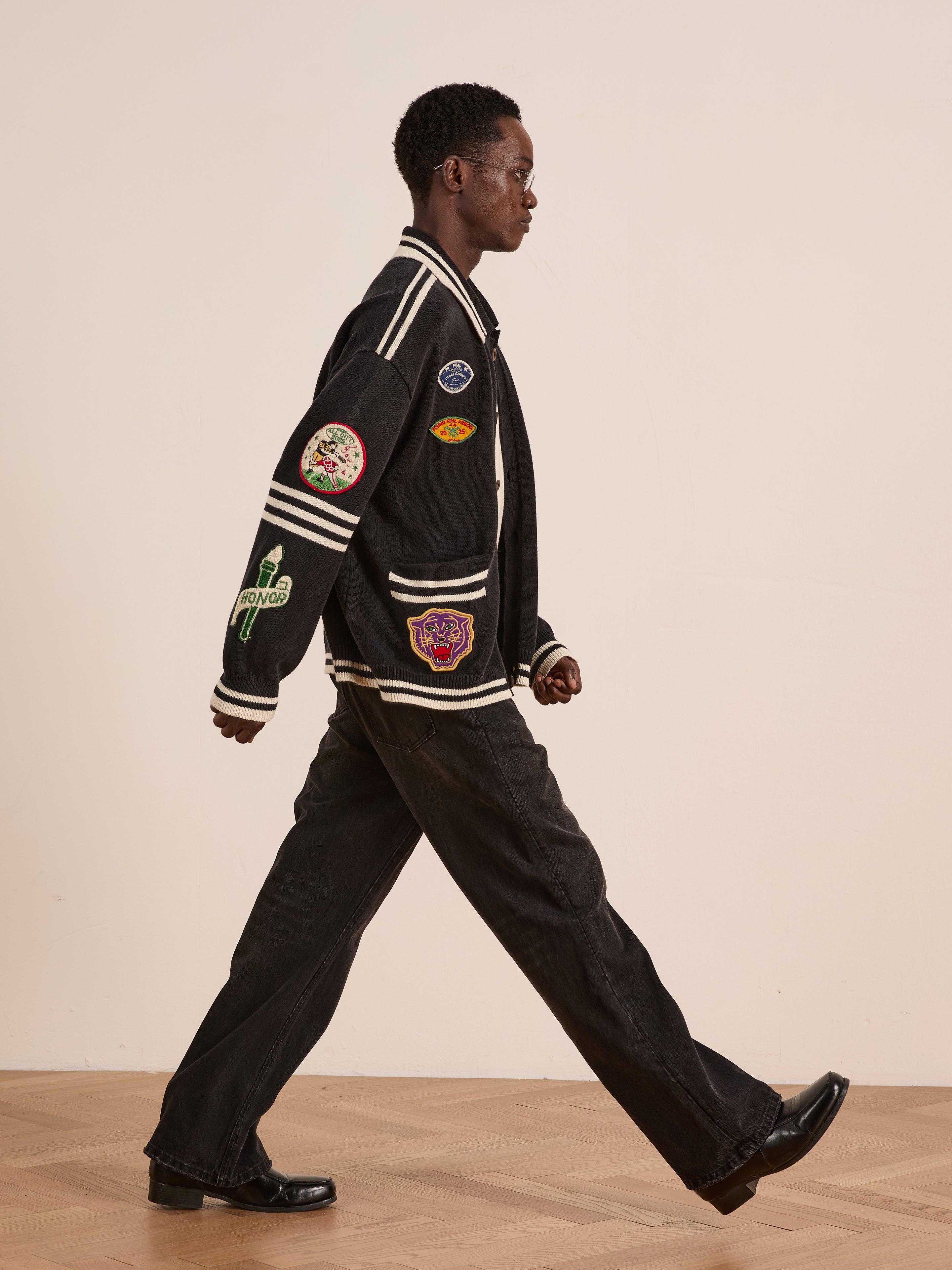 Someone wearing the FOUND Varsity Patch Collared Cardigan, paired with black pants and black shoes, walks indoors on a wooden floor against a plain background.