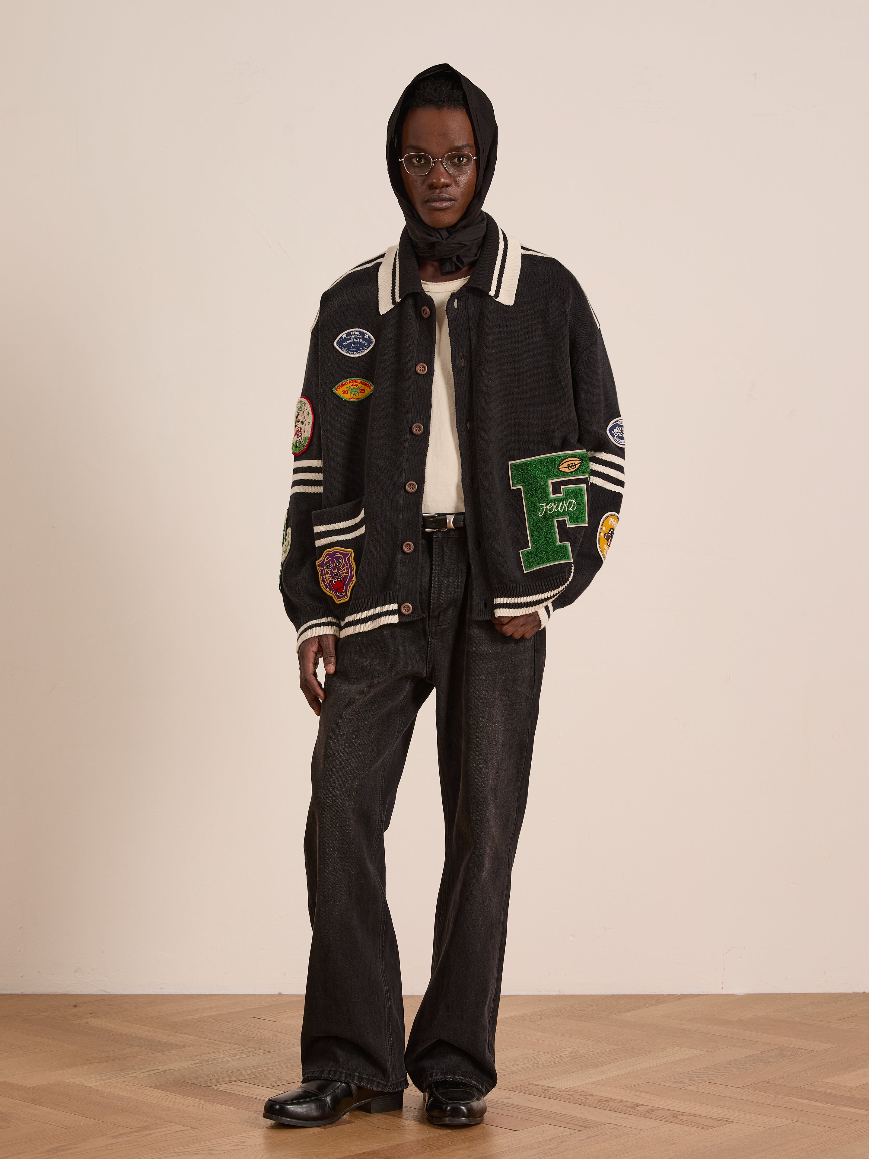 A person stands indoors on a wooden floor wearing FOUND's Varsity Patch Collared Cardigan, a black headscarf, round glasses, white shirt, black jeans, and black shoes.