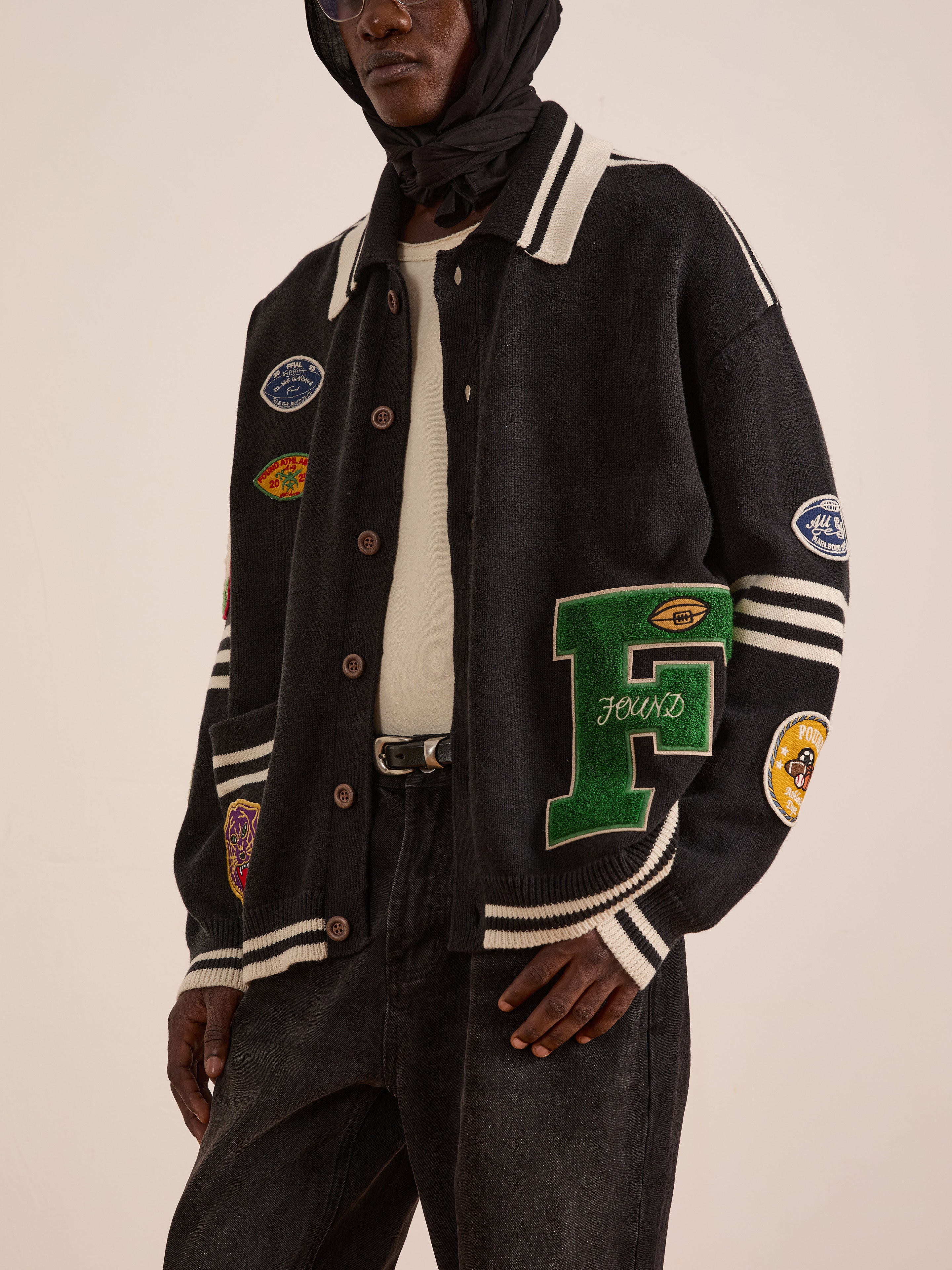 A person wearing a black FOUND Varsity Patch Collared Cardigan with patches, a white shirt, black jeans, and a headscarf stands against a plain background.