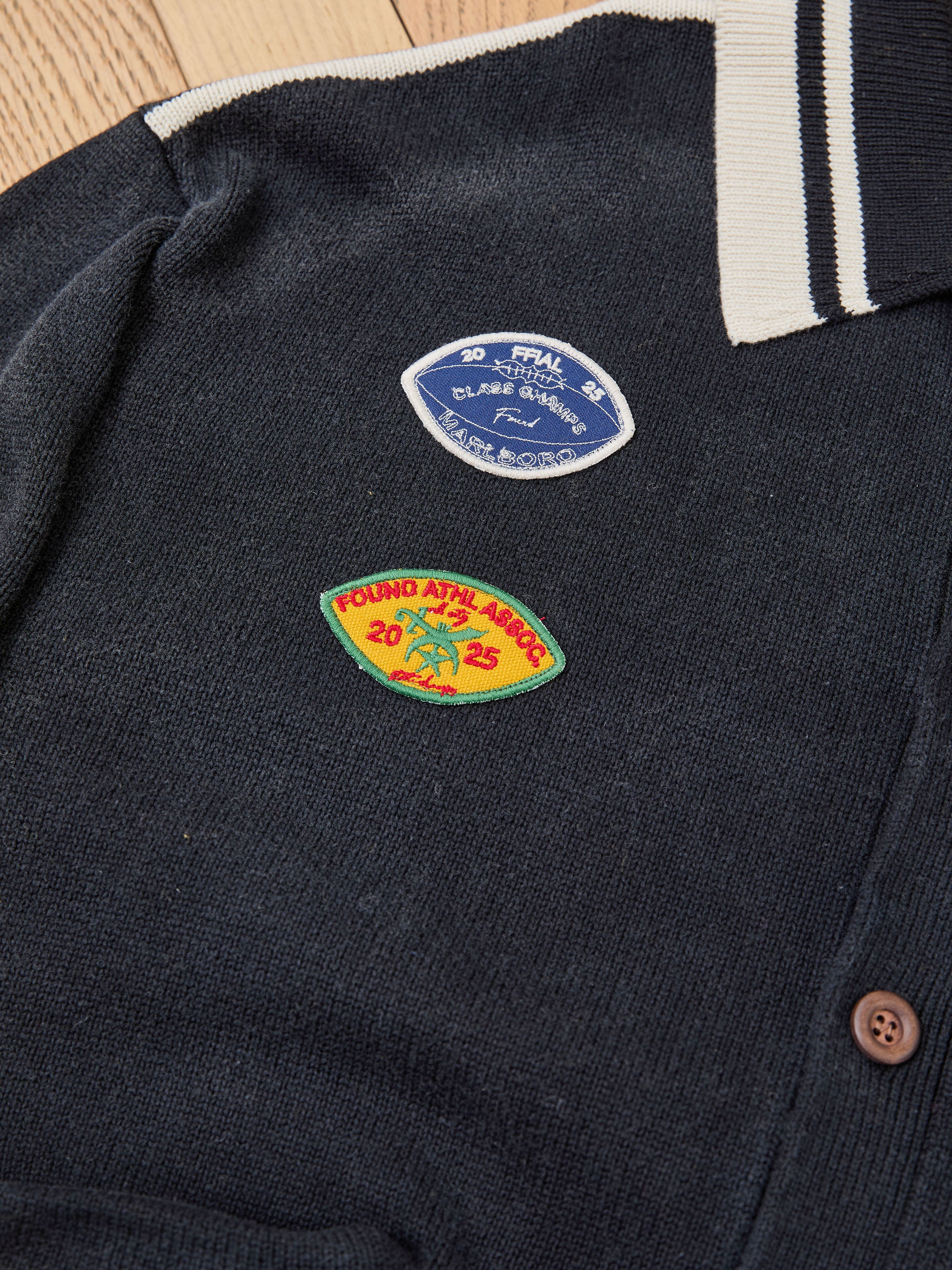 The FOUND Varsity Patch Collared Cardigan is a navy cotton knit shirt with a white-trimmed collar, featuring two chenille chest patches—a blue oval and a yellow-green shield—each with unique text and graphics.