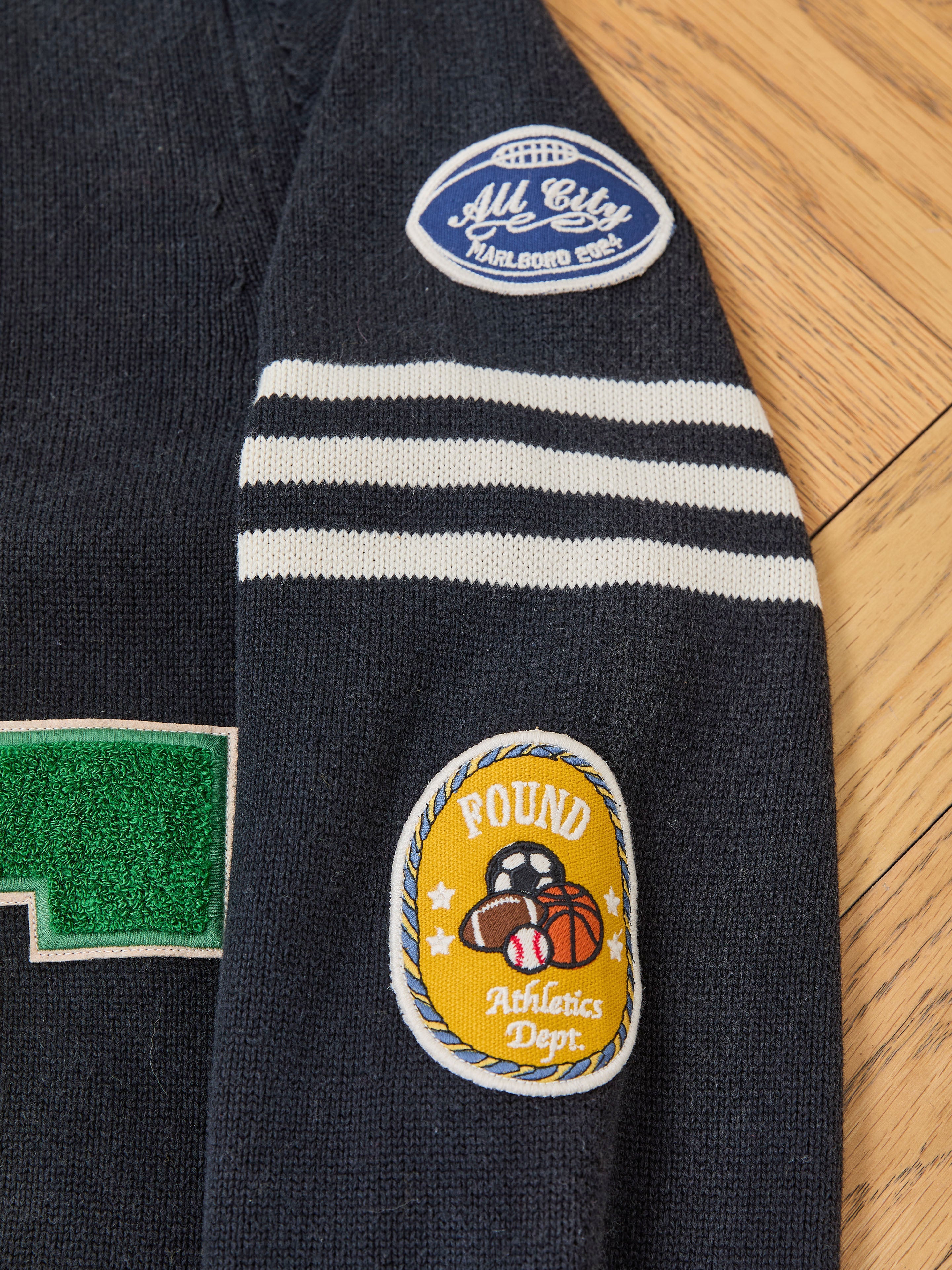 The FOUND Varsity Patch Collared Cardigan in navy cotton knit features white stripes and two chenille patches: an oval blue "All City Karlborg Edge" patch and a yellow "Pound Athletics Dept." patch with sports ball graphics.