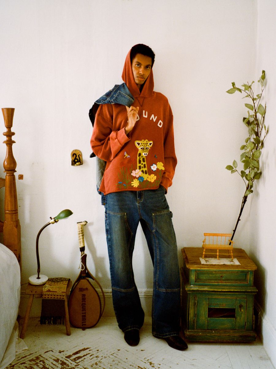 In a room, someone wears a red hoodie featuring a giraffe design paired with FOUND Denim Carpenter Jeans, casually draping a denim jacket over their shoulder. Nearby stands a green table adorned with a plant, next to both a lamp and bed.
